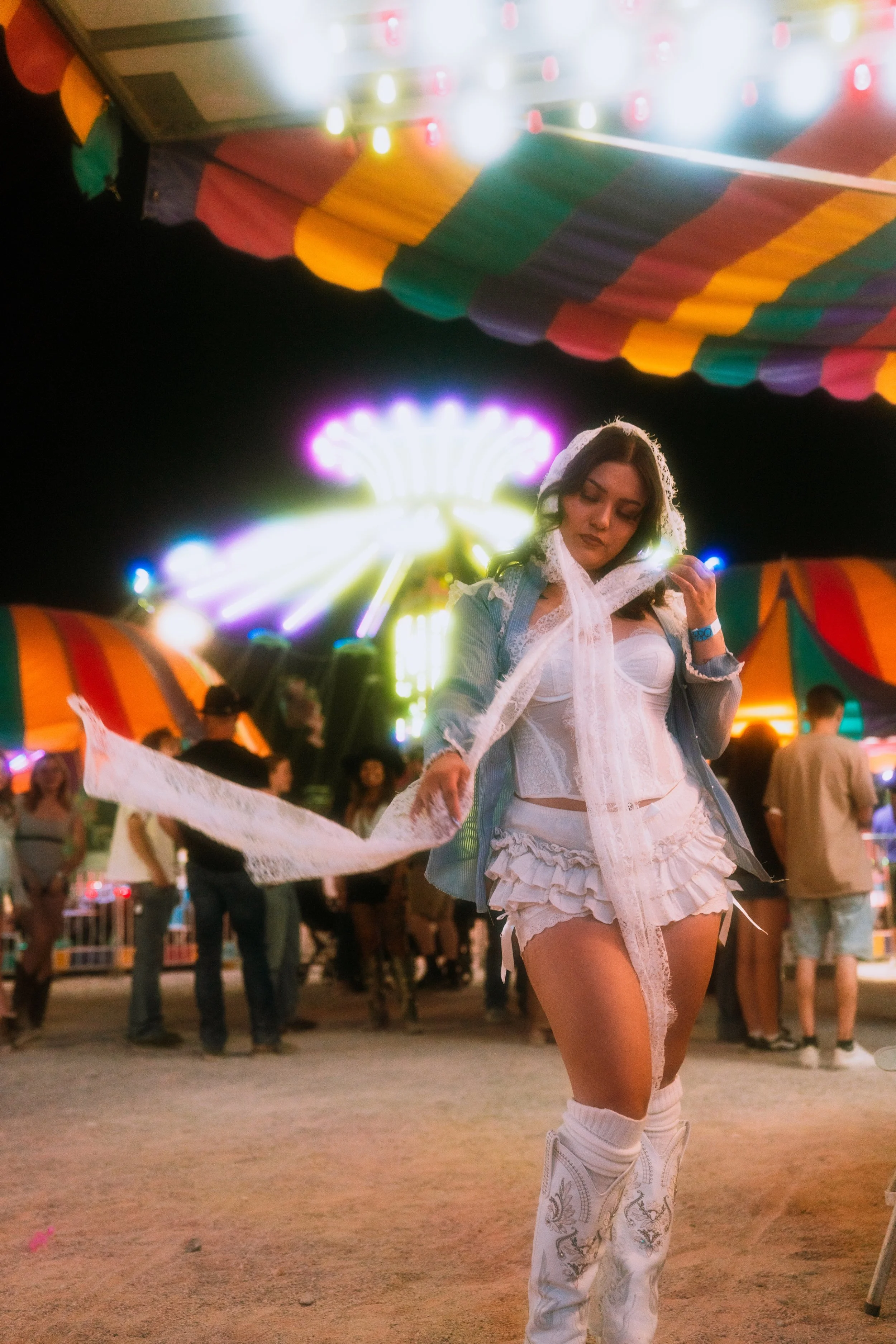 A young woman dressed in white lace attire, standing at a nighttime carnival or fair. She is wearing a lace hood, a white bustier top, ruffled shorts, knee-high socks, and embroidered cowboy boots. Bright colorful tents and a lit-up amusement ride ar