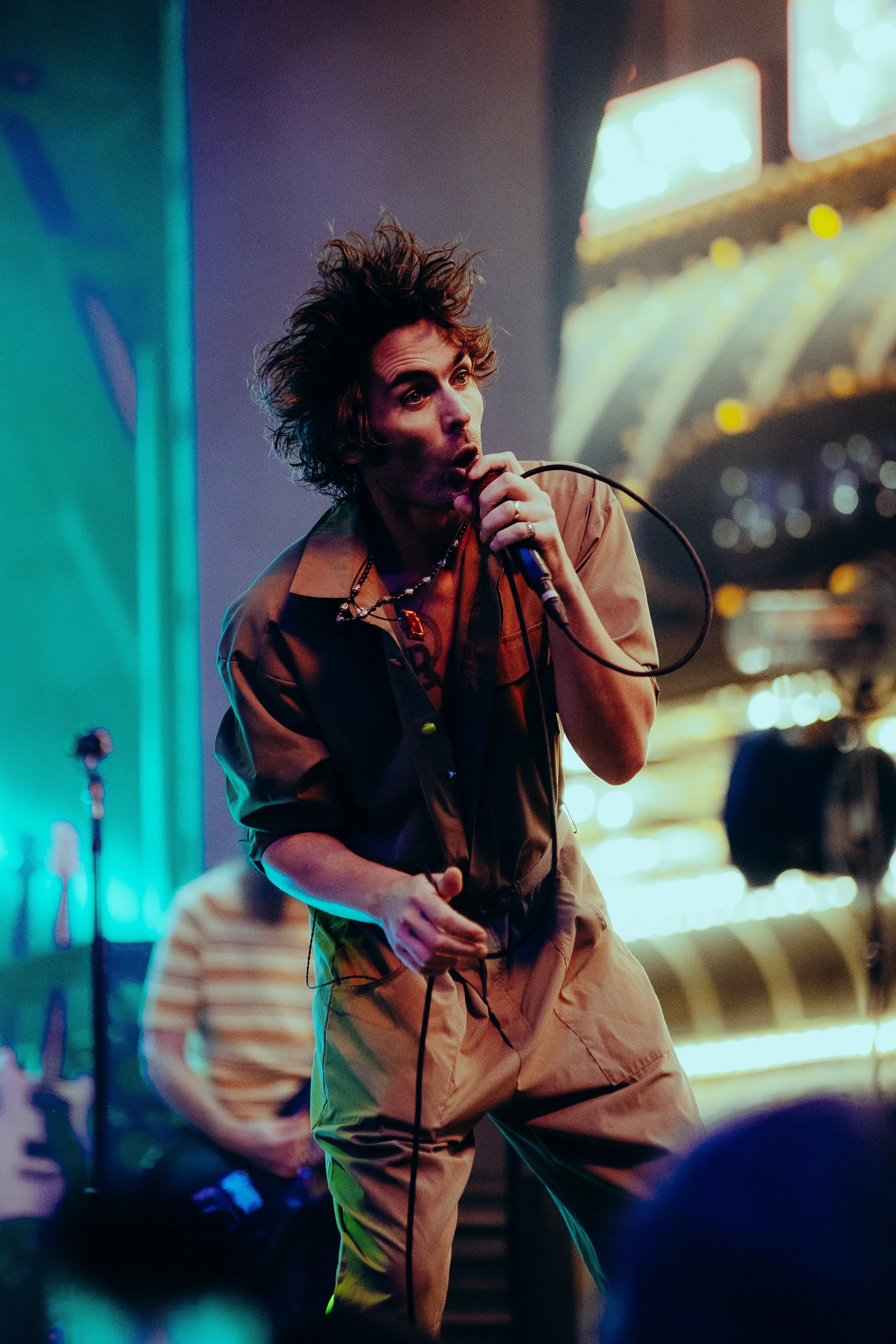 A person with curly hair singing into a microphone, wearing a tan jacket and pants, with colorful lights in the background.
