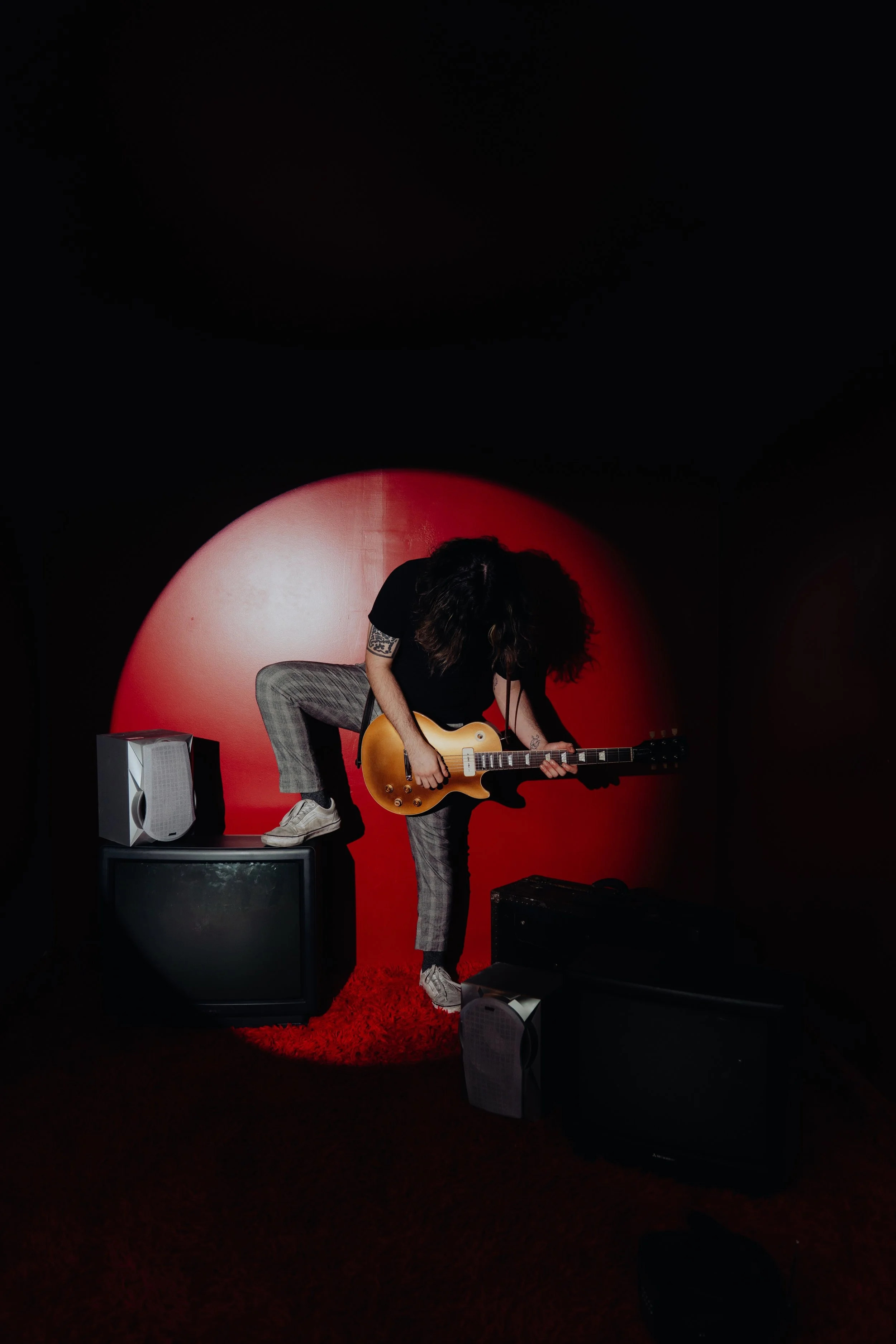 A person with long curly hair, wearing a black t-shirt and plaid pants, is playing an electric guitar on a small stage with a red background. The person is standing on a red rug and is surrounded by amplifiers and speakers.