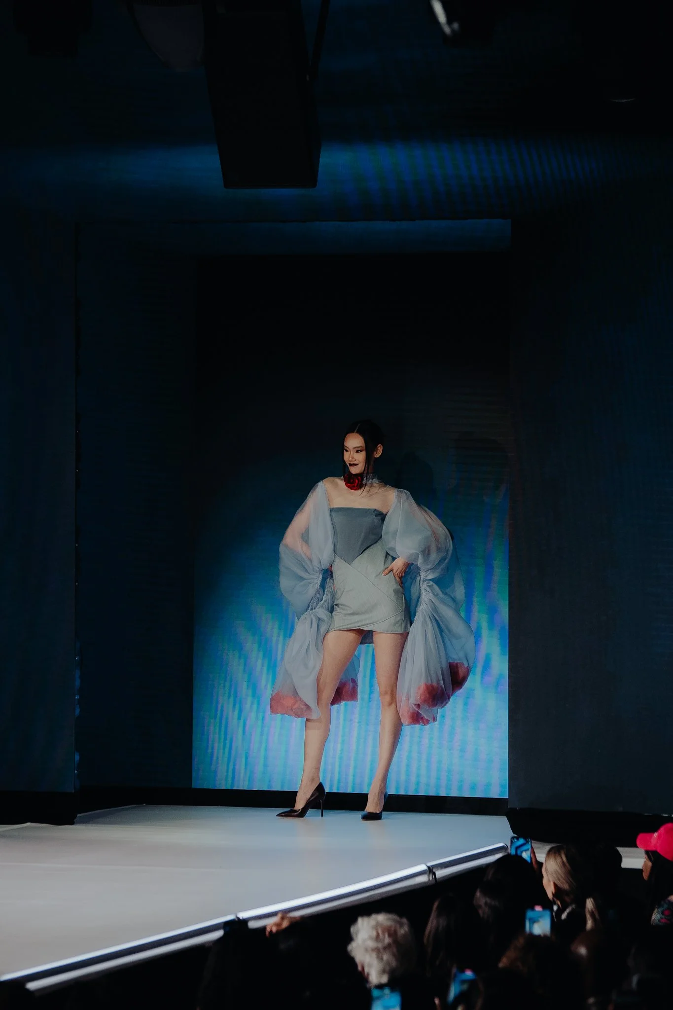 A female model on a runway wears a light-colored dress with sheer, puffy sleeves and a floral design, paired with black high heels, in front of a blue digital background and an audience watching.