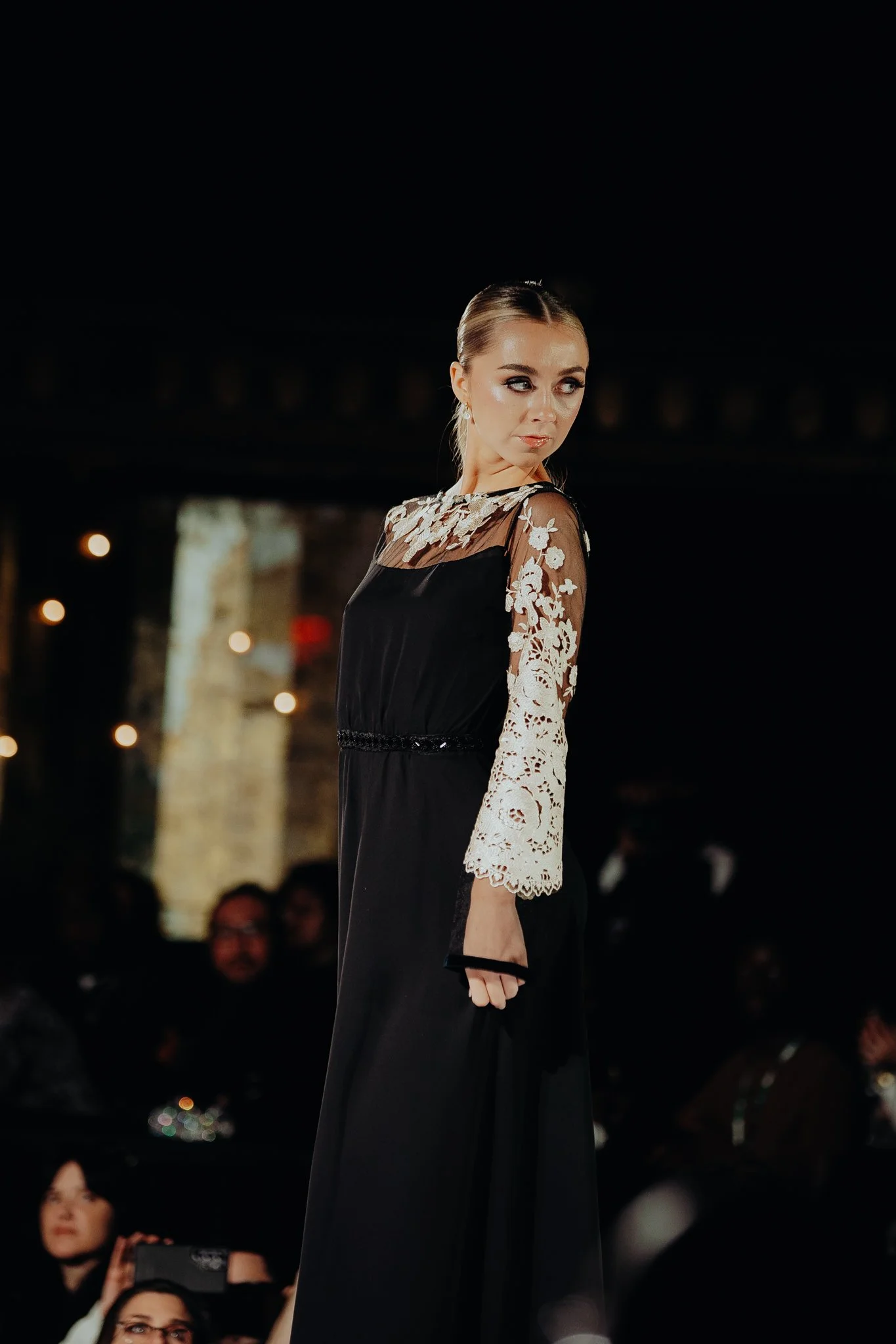 A woman in a black dress with white lace sleeves walking on a runway during a fashion show with a dark background and blurred audience.