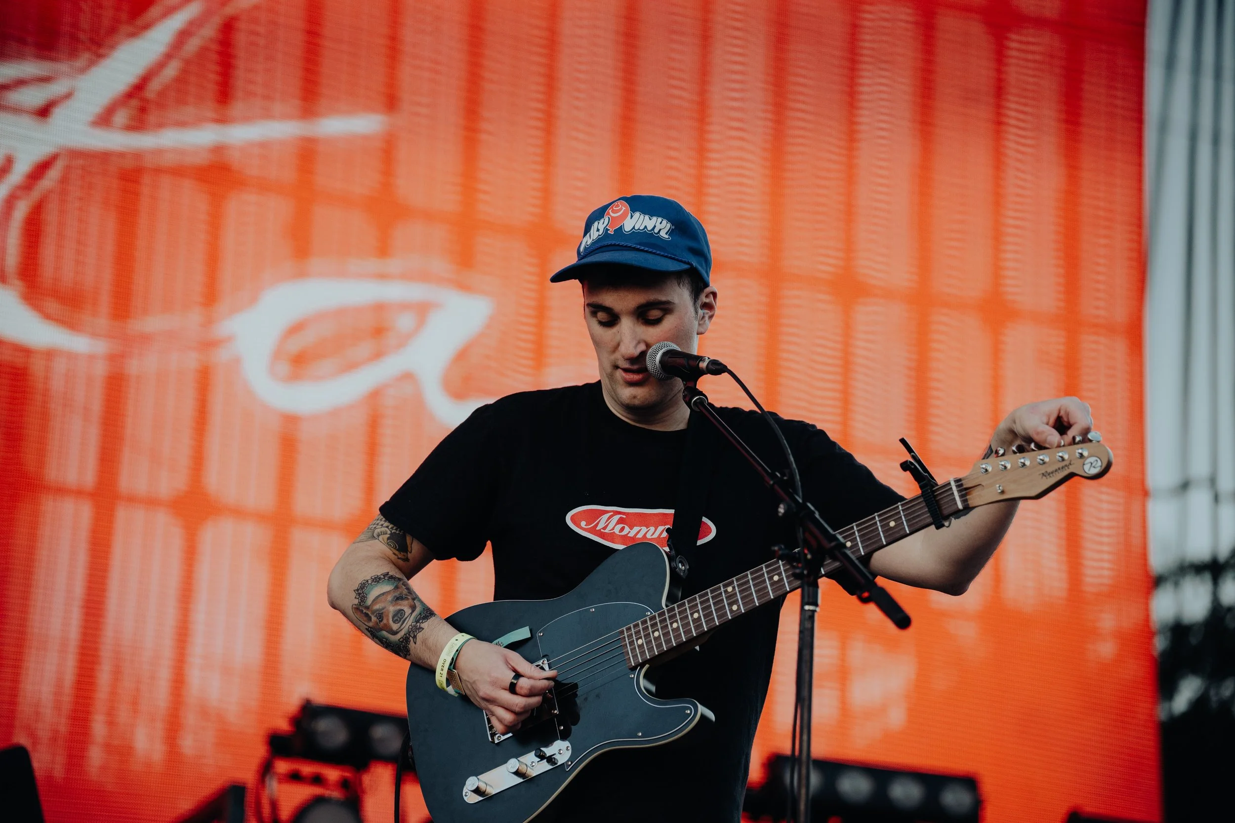 A young man with tattoos on his arm wearing a black T-shirt and a blue baseball cap playing a black electric guitar on stage with an orange background.