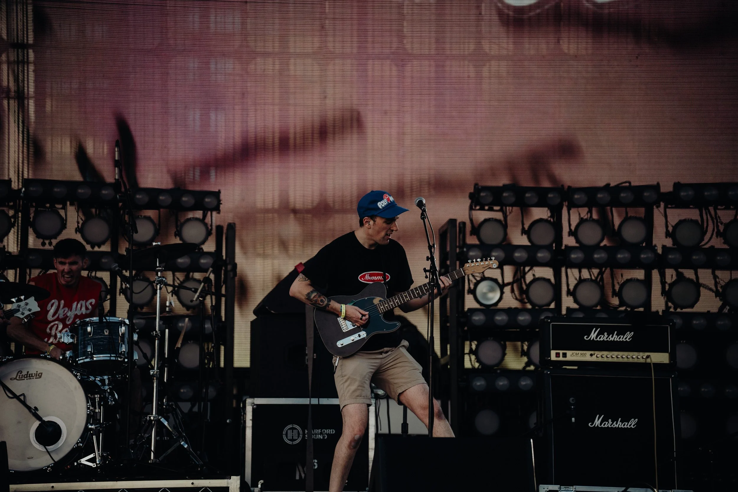 A band performing live on stage with musical instruments, including a drum set and electric guitars, in front of a large screen showing a close-up of a face.