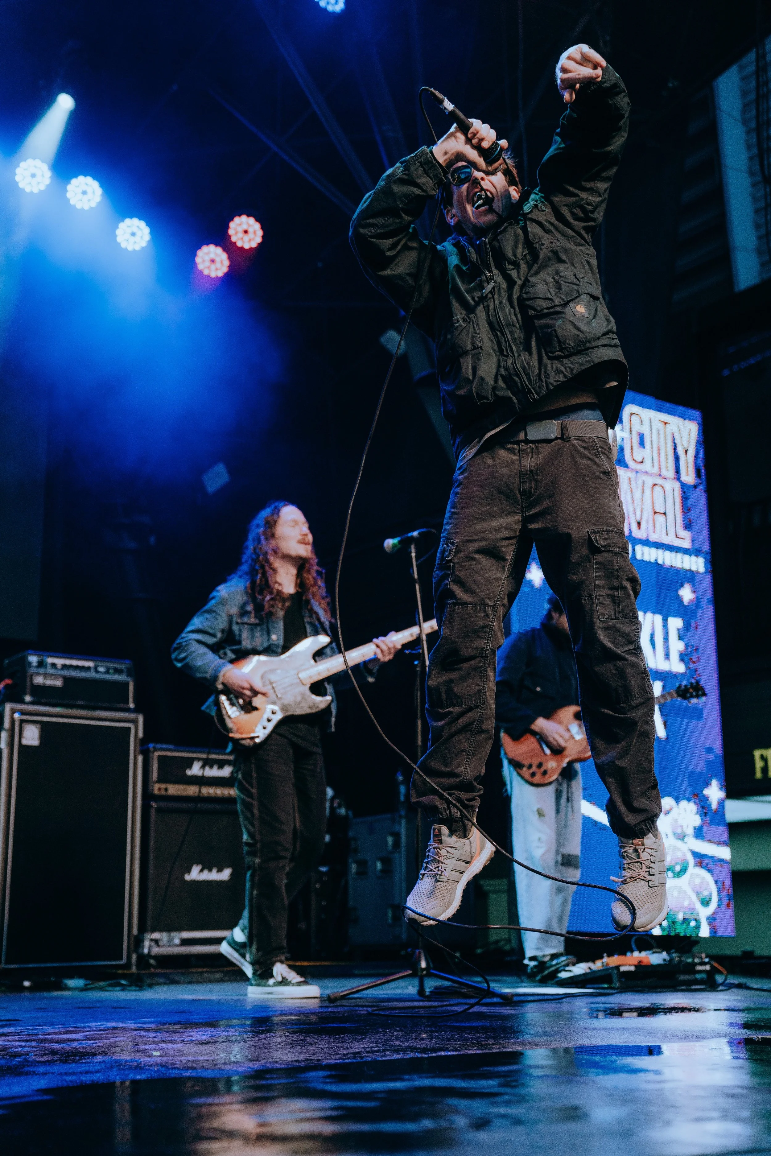A performer singing on stage with a microphone, wearing a black jacket, cargo pants, and white sneakers, with a band playing instruments behind him, including a guitarist and a bassist, at a concert event.