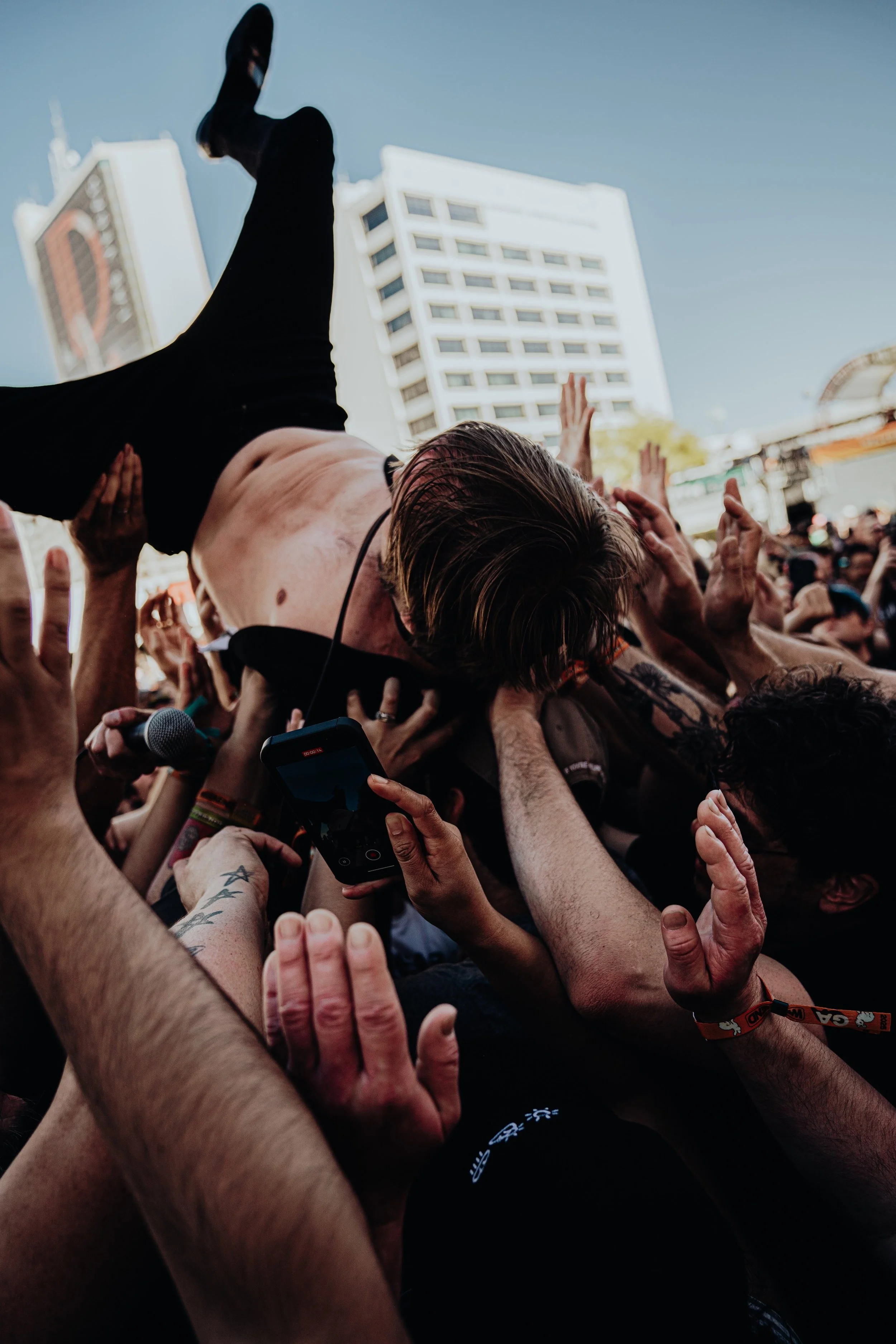 A crowd of people at an outdoor concert or event, crowd surfing with a person being lifted above the crowd, in front of buildings under a clear blue sky.