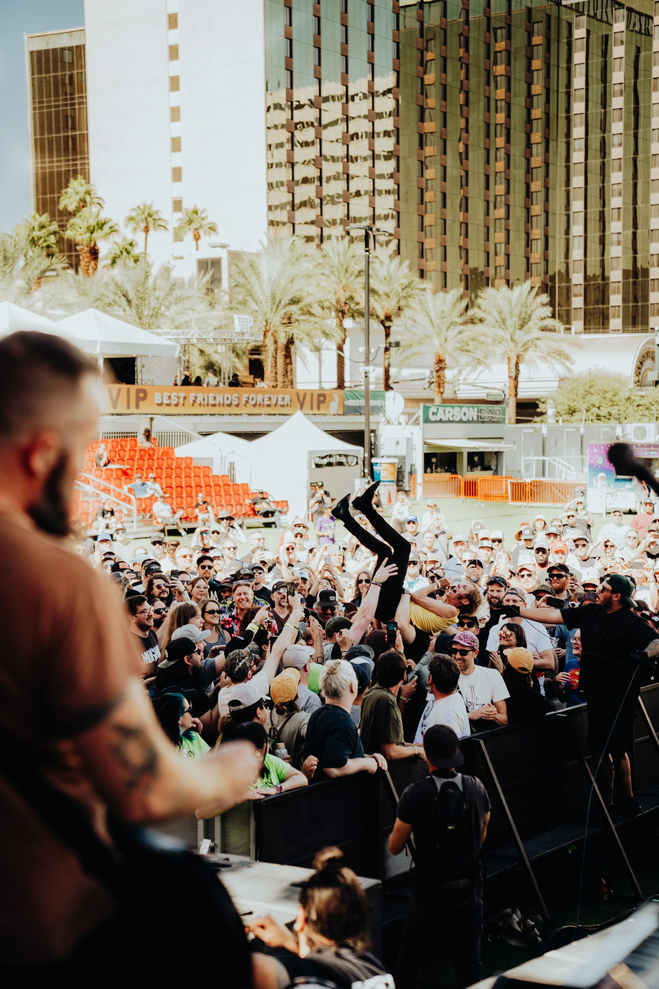 Crowd of people attending an outdoor concert or festival with a stage in the background and tall modern buildings, palm trees, and tents.