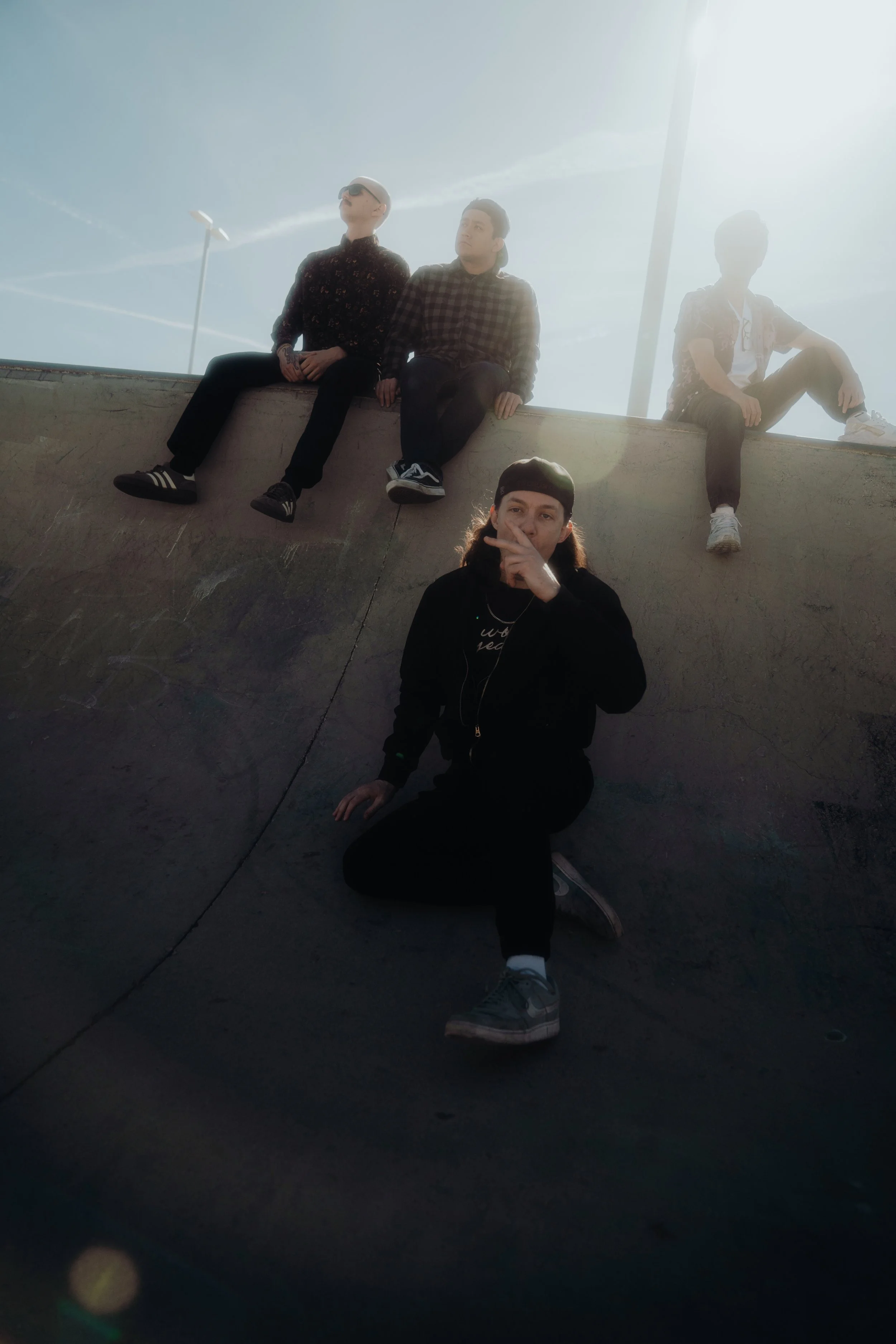 Four young men sitting on a concrete ledge outdoors with bright sunlight and a blue sky, some wearing sunglasses and casual clothing, one making a peace sign gesture.
