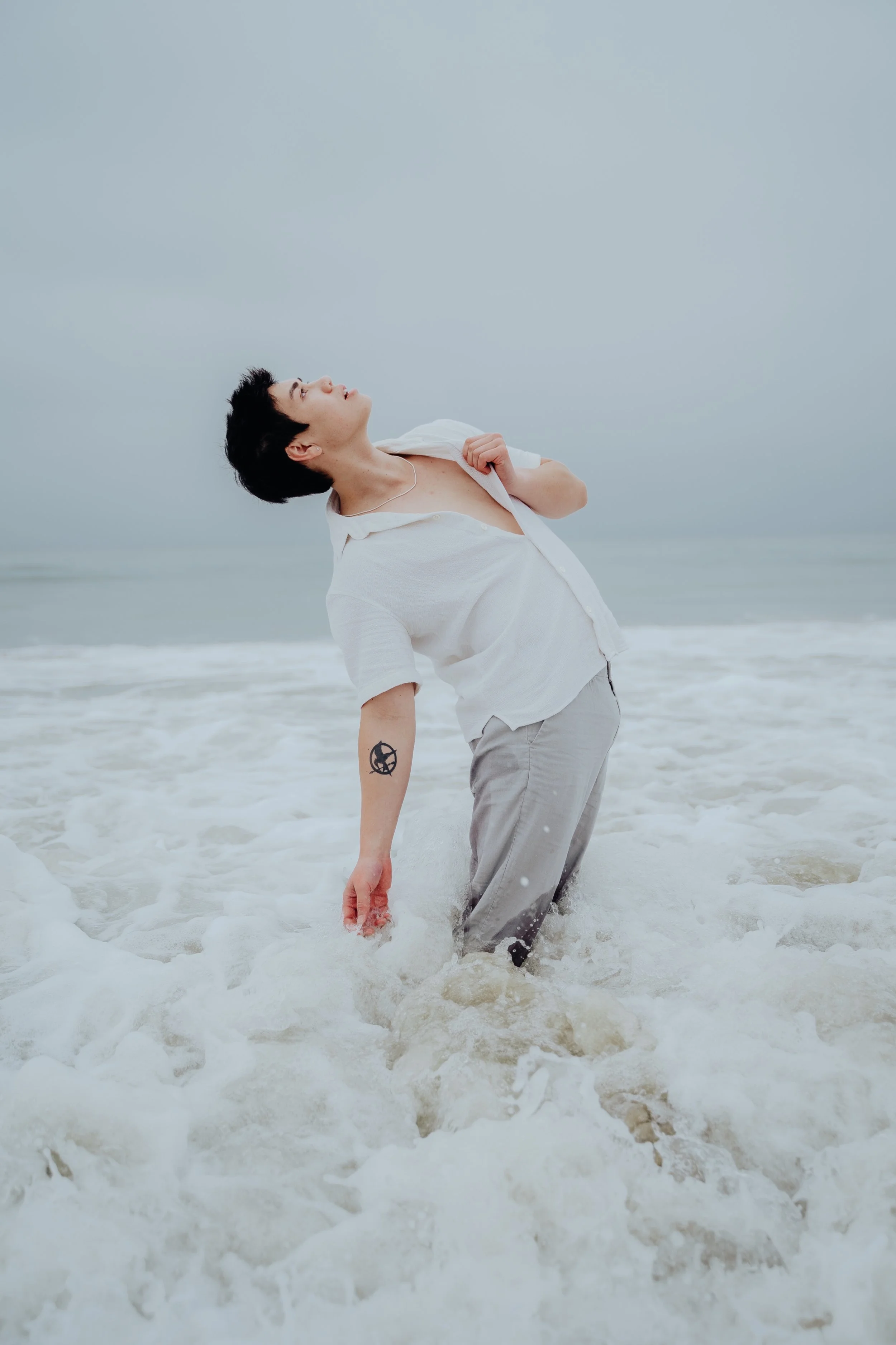 Person standing in the ocean with waves reaching their knees, wearing a white shirt and Light-colored pants with tattoos on their left arm, looking upwards with a cloudy sky in the background.