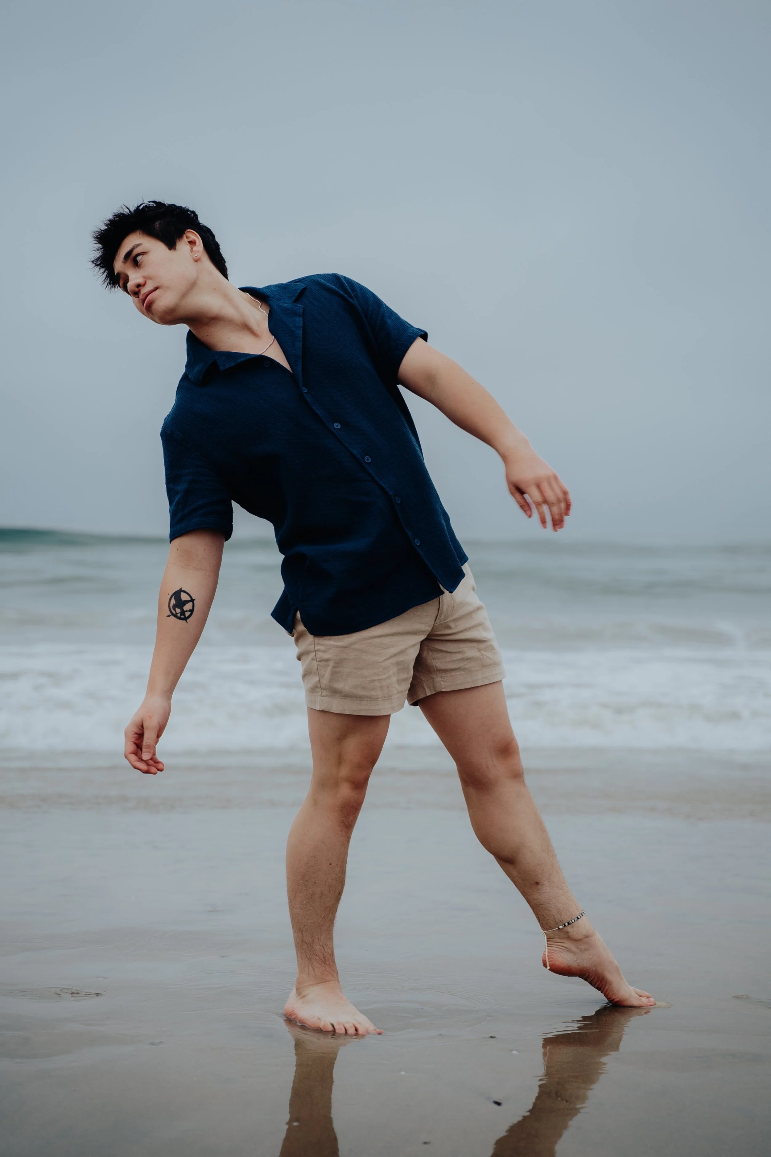 A young man in a dark blue shirt and beige shorts standing barefoot on the wet sand at the beach, leaning to his left with a tattoo on his left forearm, and a thin ankle bracelet on his right ankle, during daytime with an overcast sky.
