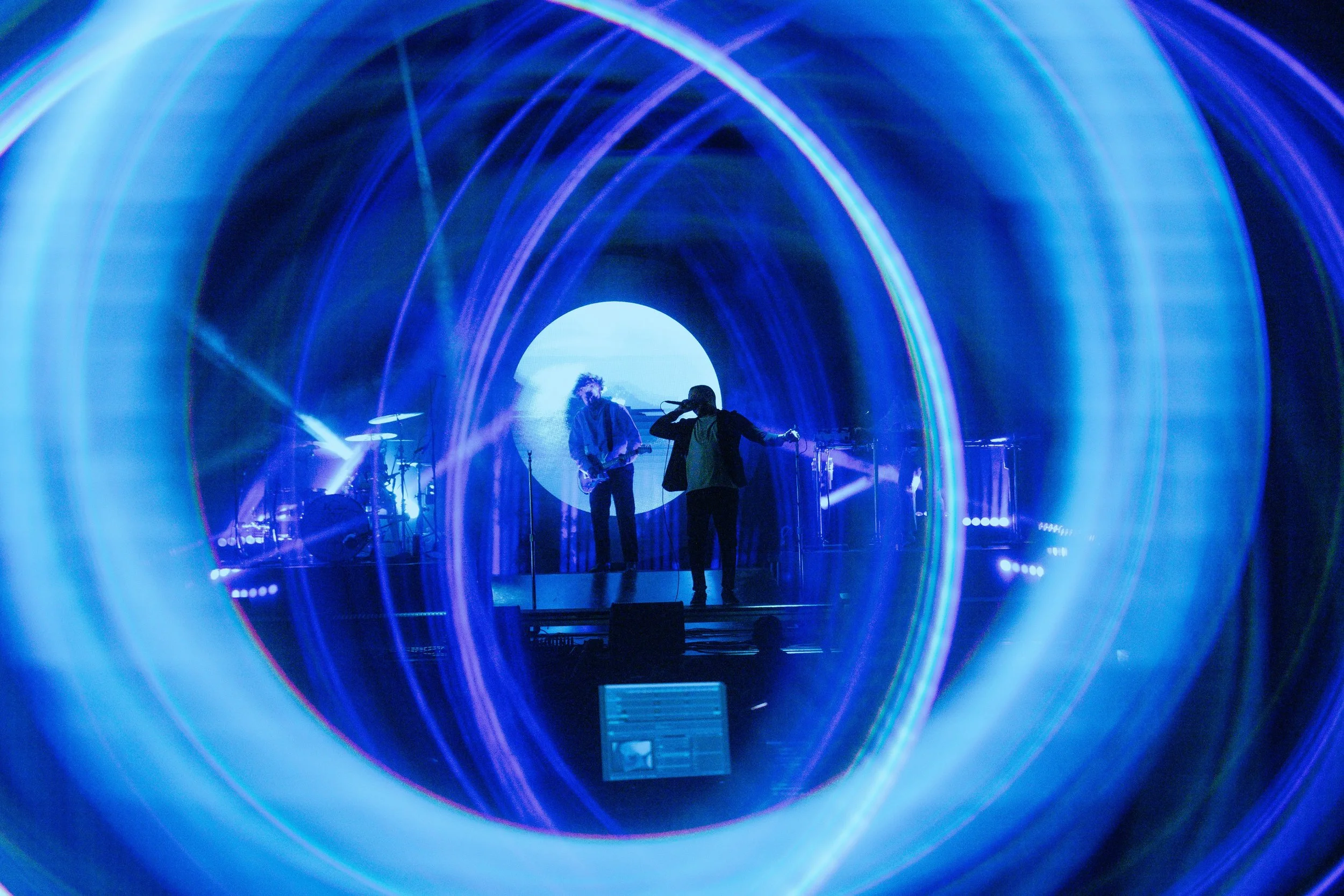 A live performance with two musicians on stage framed by a circular light tunnel effect, with vibrant blue and purple light beams and a scenic background.