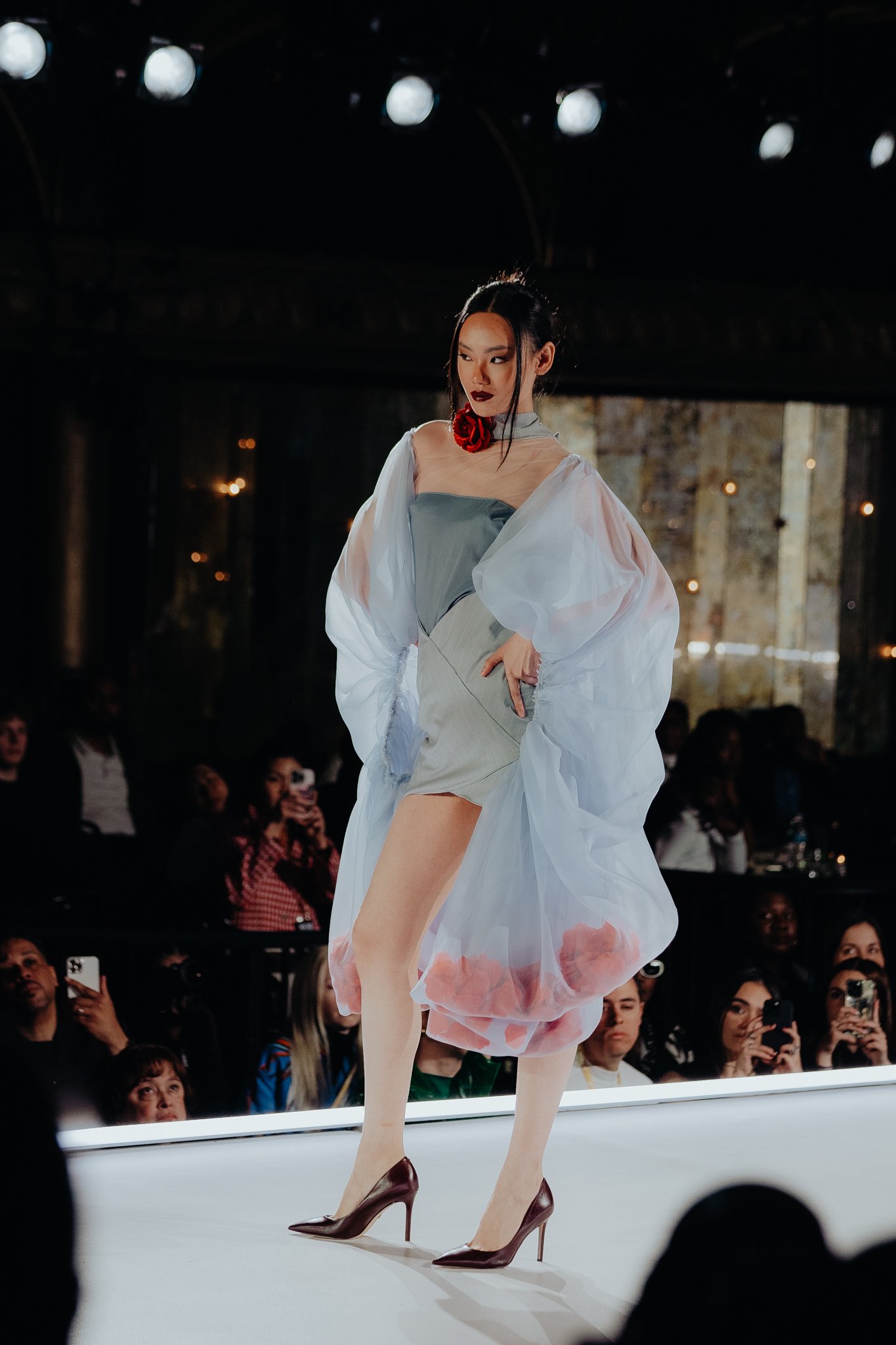 Fashion model walking on runway during fashion show, wearing dress with sheer top, floral accents, and high heels, with audience watching.