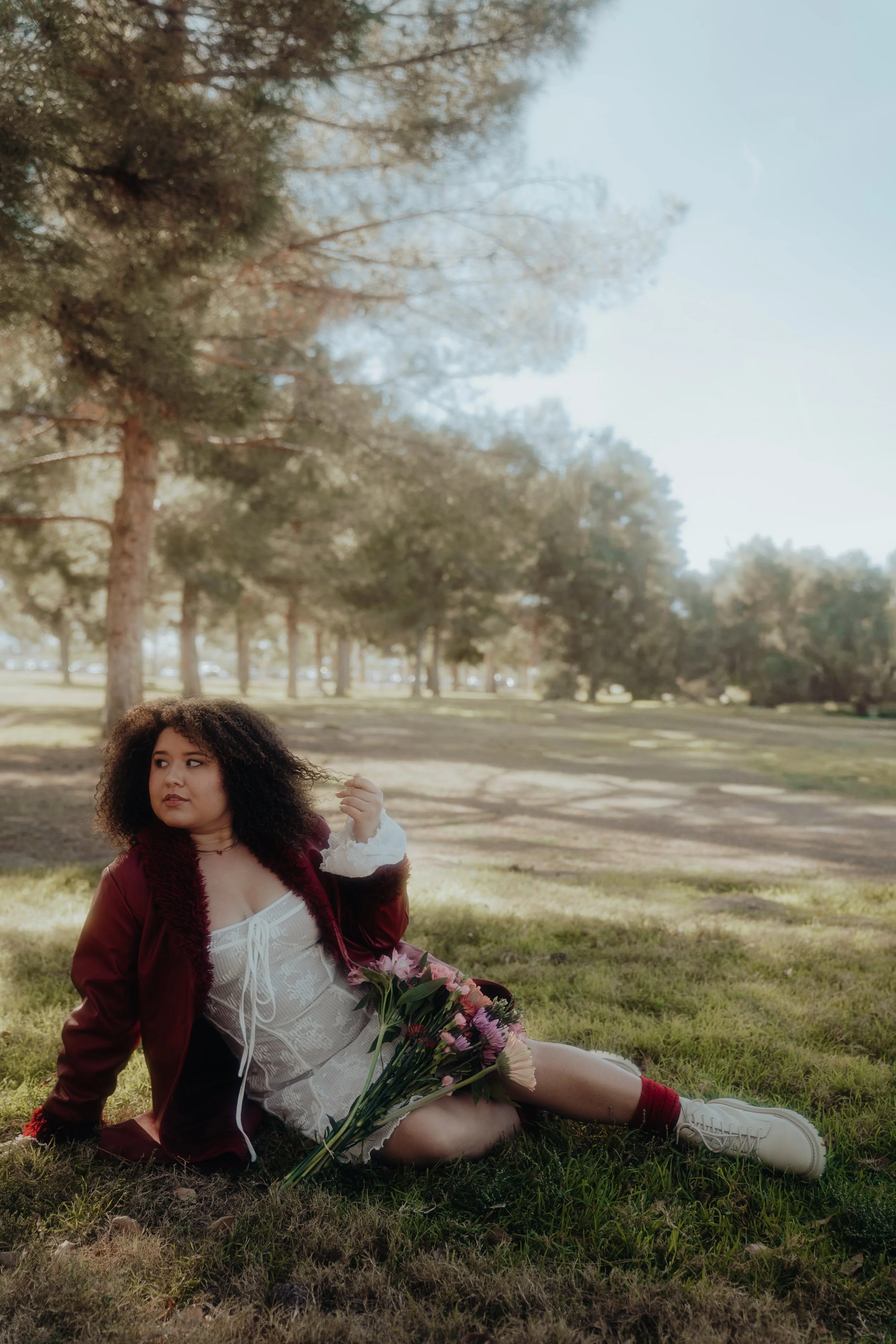 A woman with curly hair sitting on grass in a park, holding a bouquet of flowers, surrounded by trees and sunlight.