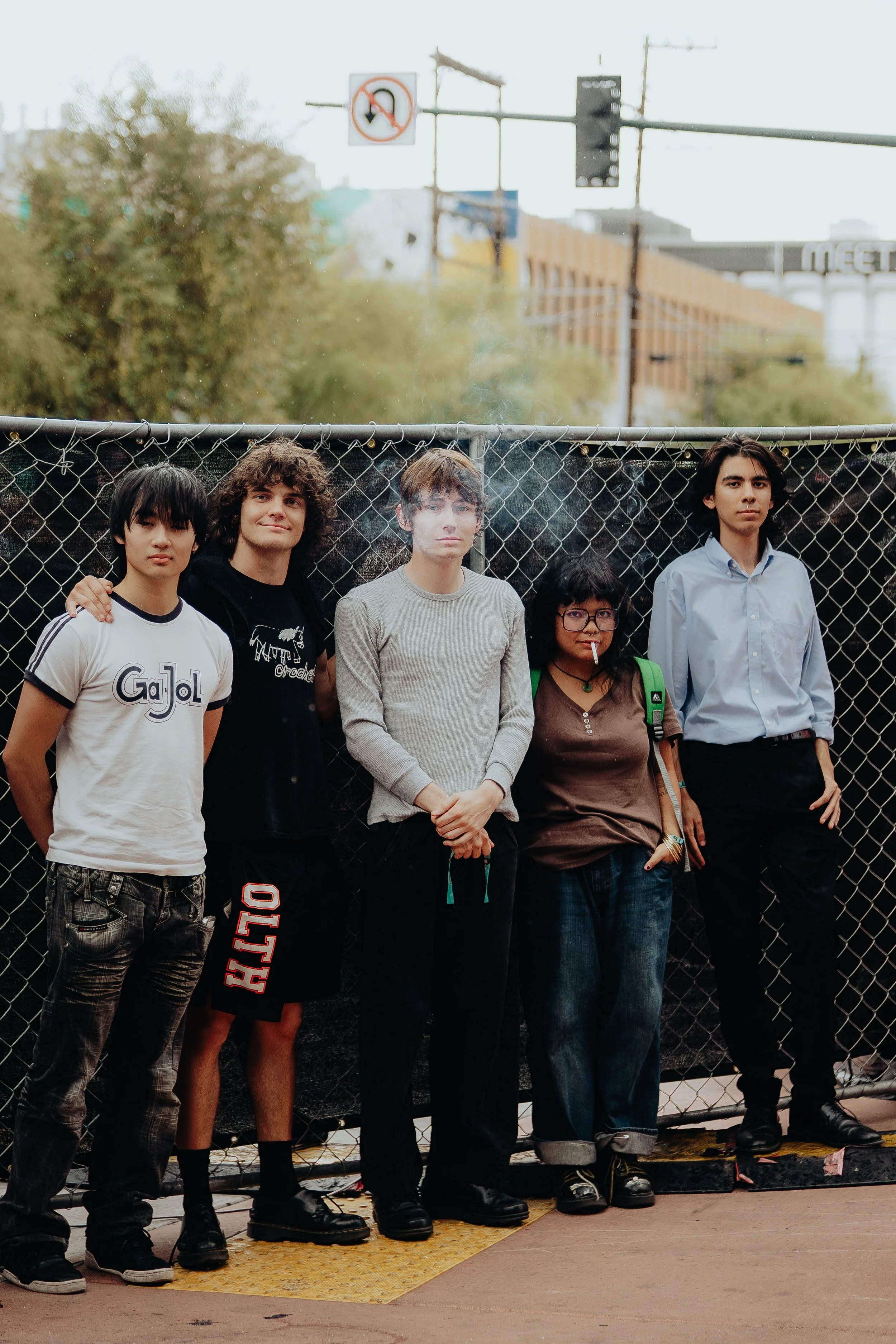Group of five young adults standing side by side behind a chain-link fence, with city buildings and traffic signals in the background.