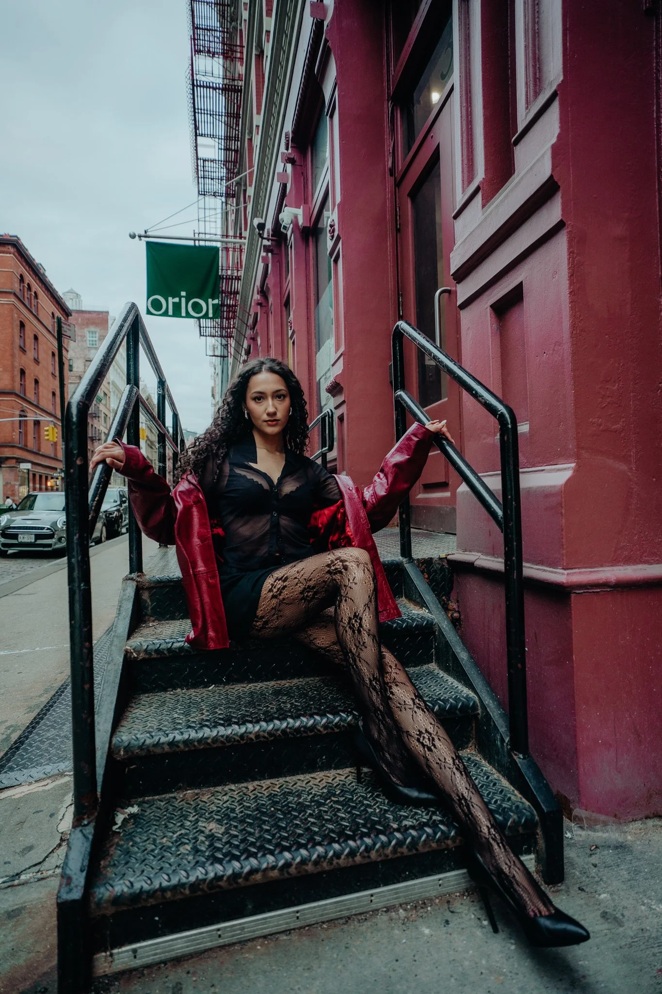 A woman sitting on metal stairs outside a red building on an urban street, with brick buildings and parked cars in the background, holding onto the stair railing.