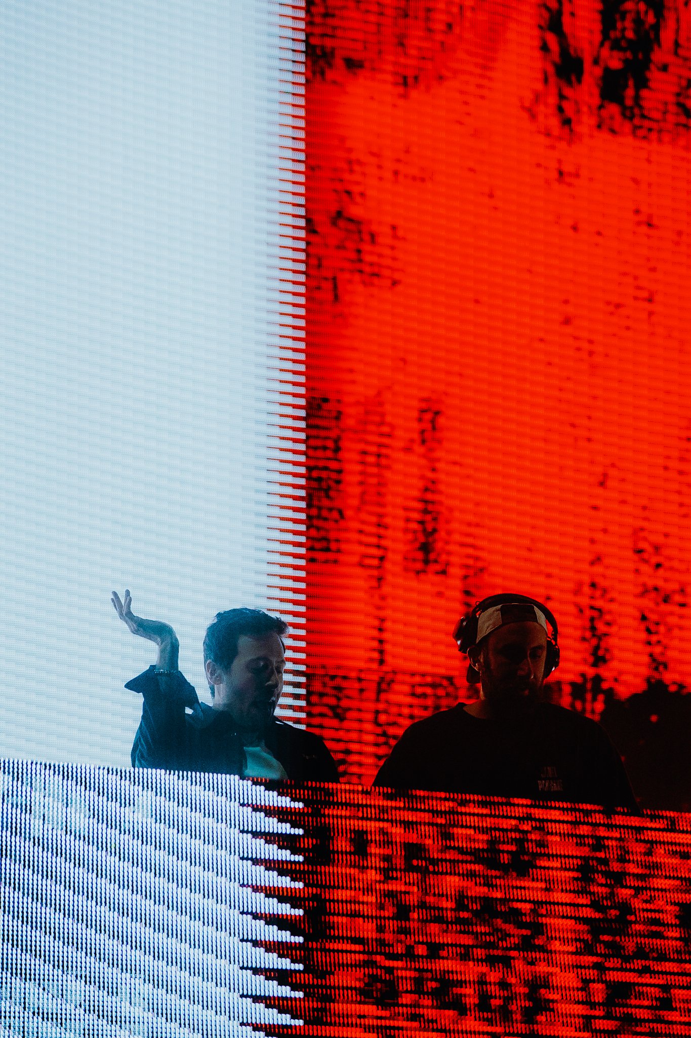 Two individuals standing in front of a large digital screen displaying a mix of red, black, and white colors with pixelated patterns. One person appears to be speaking or gesturing, while the other person is facing forward and wearing a cap.
