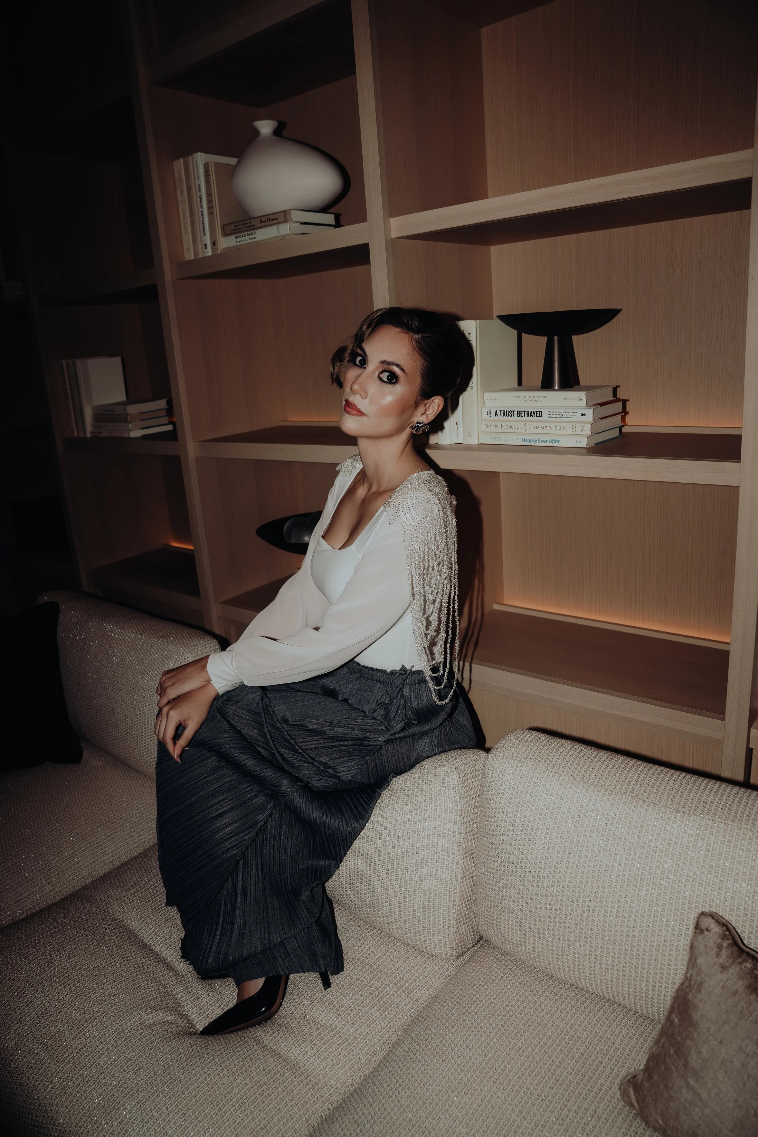 A woman with short dark hair, wearing a white blouse and dark pleated skirt, is sitting on a beige sofa in a living room with wooden bookshelves behind her. The shelves contain books and decorative objects, including a white vase and a black bowl. Th