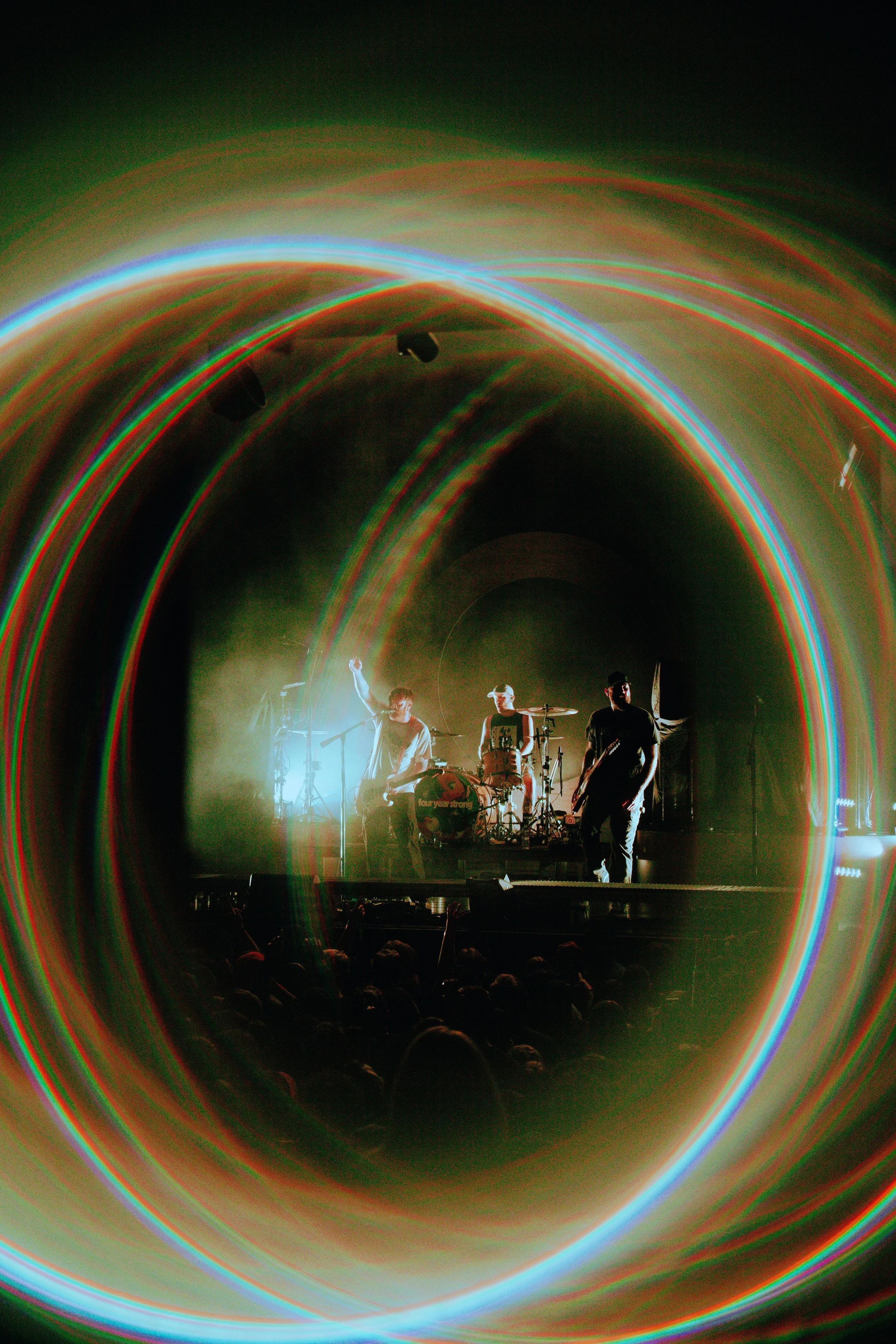 A live music performance on stage with four band members, colorful light trails arcing above and around them, creating a dynamic and vibrant atmosphere.