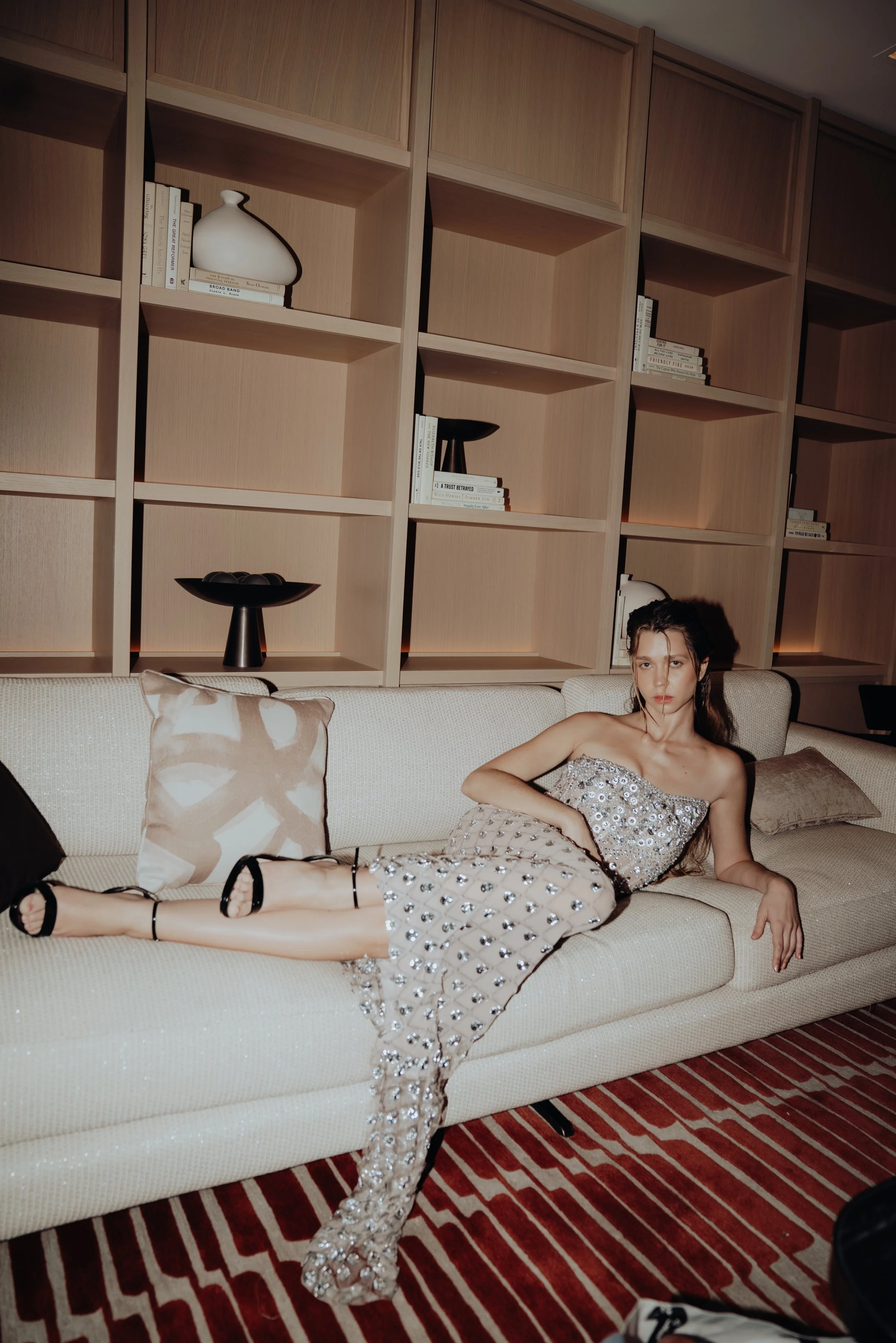 A young woman with dark hair sitting on a beige couch in a room with wooden shelves and books behind her. She is wearing a strapless, sequined dress with a slit, black strappy high heels, and has her legs crossed. The room has a patterned red and bei