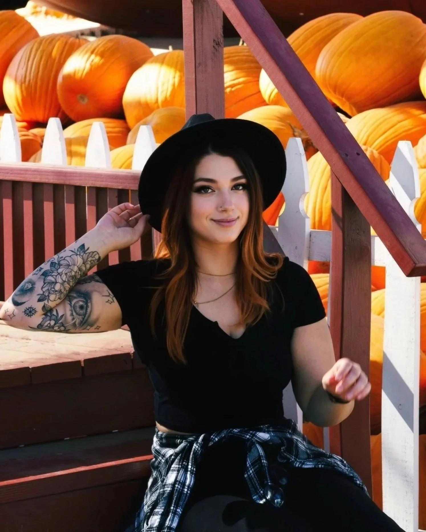 A young woman with tattoos on her arm, wearing a black hat and black shirt, sitting outdoors in front of a display of large orange pumpkins, with a wooden fence and stairs in the background.