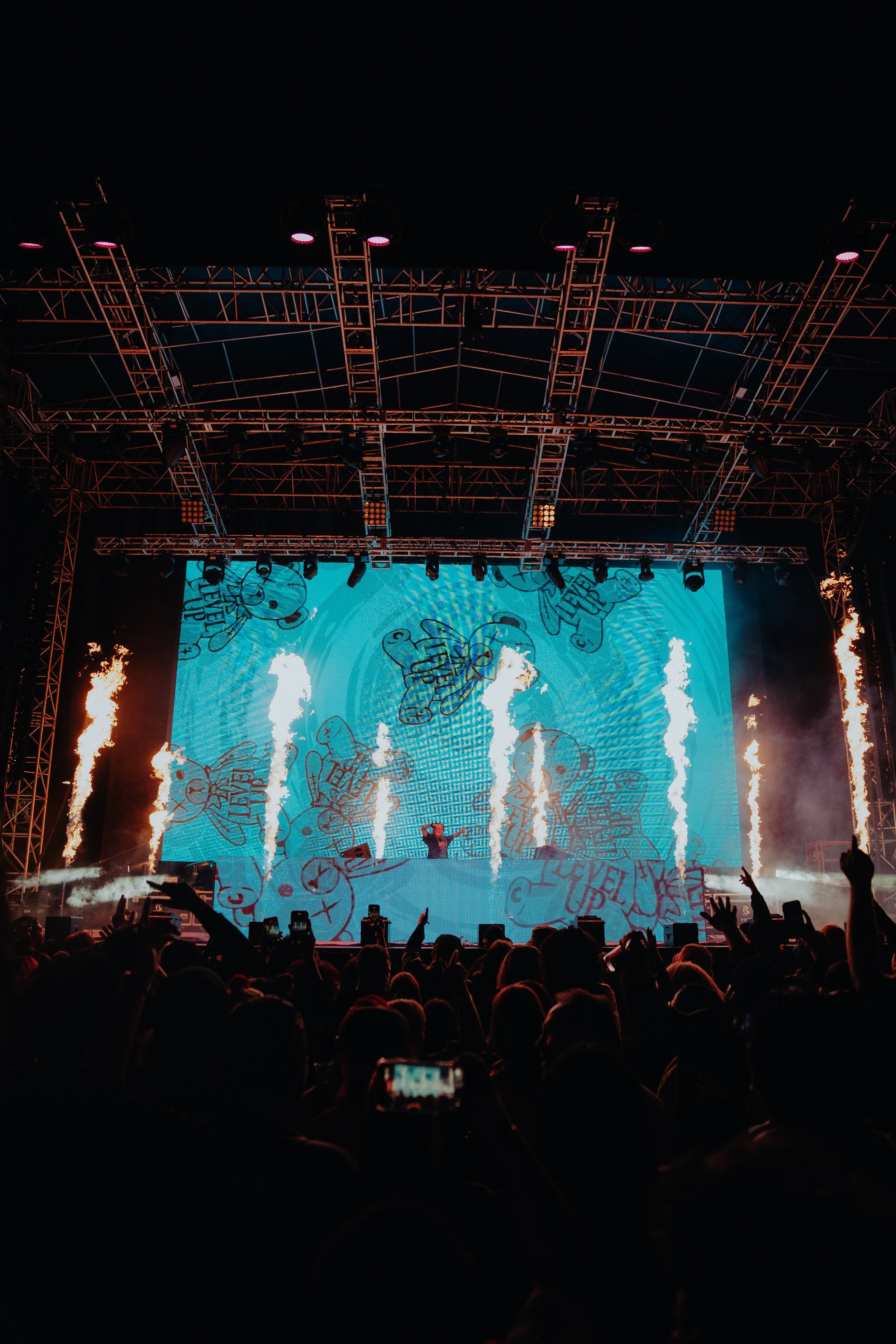 A large crowd at a concert or festival viewing a stage with a digital screen displaying cartoon characters with speech bubbles, flames rising at the sides, and a DJ or performer in front of the screen, with audience members raising their hands.