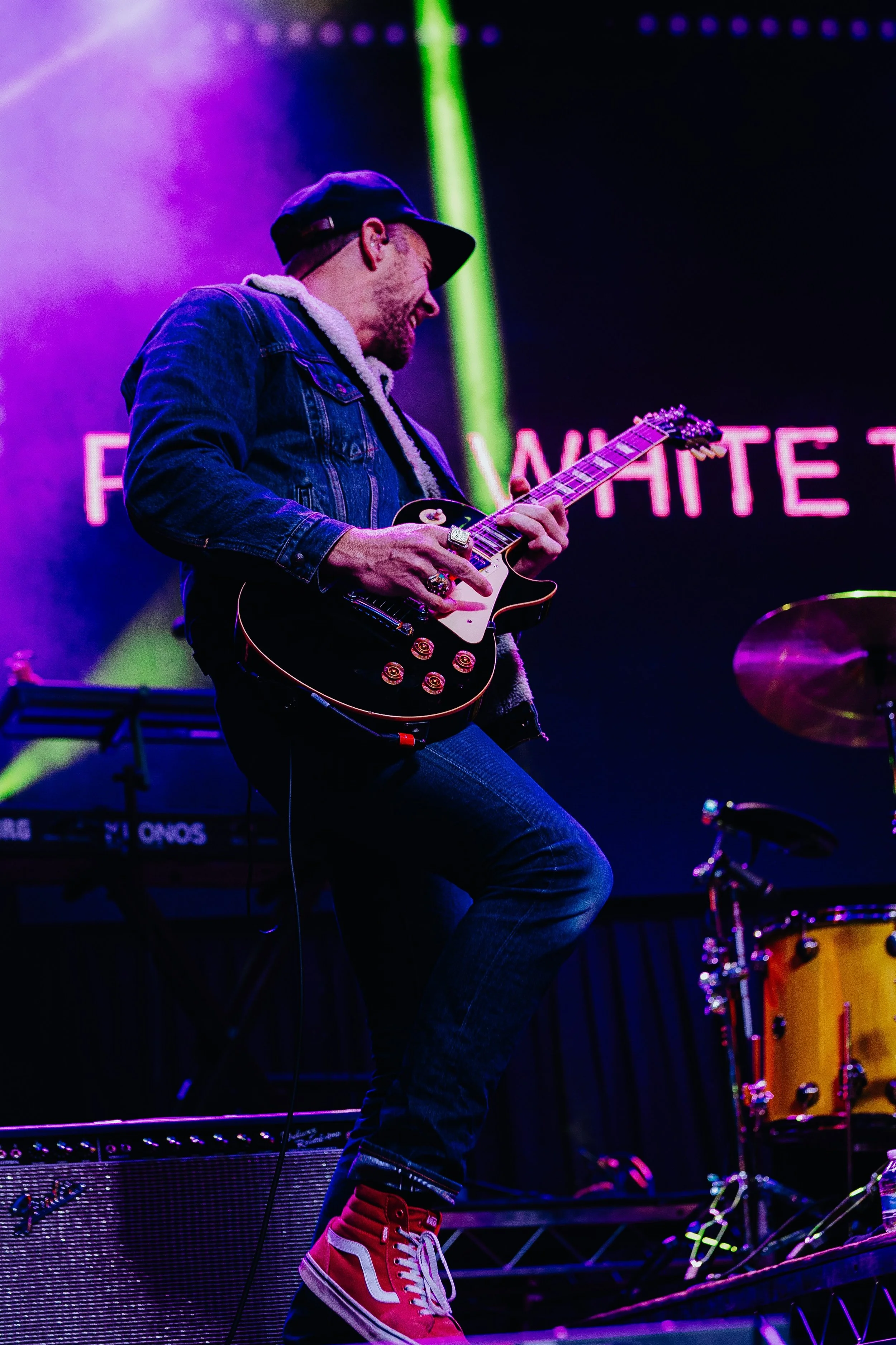 Musician playing electric guitar on stage with vibrant neon purple and green lights, wearing a black cap, denim jacket, and red high-top sneakers.