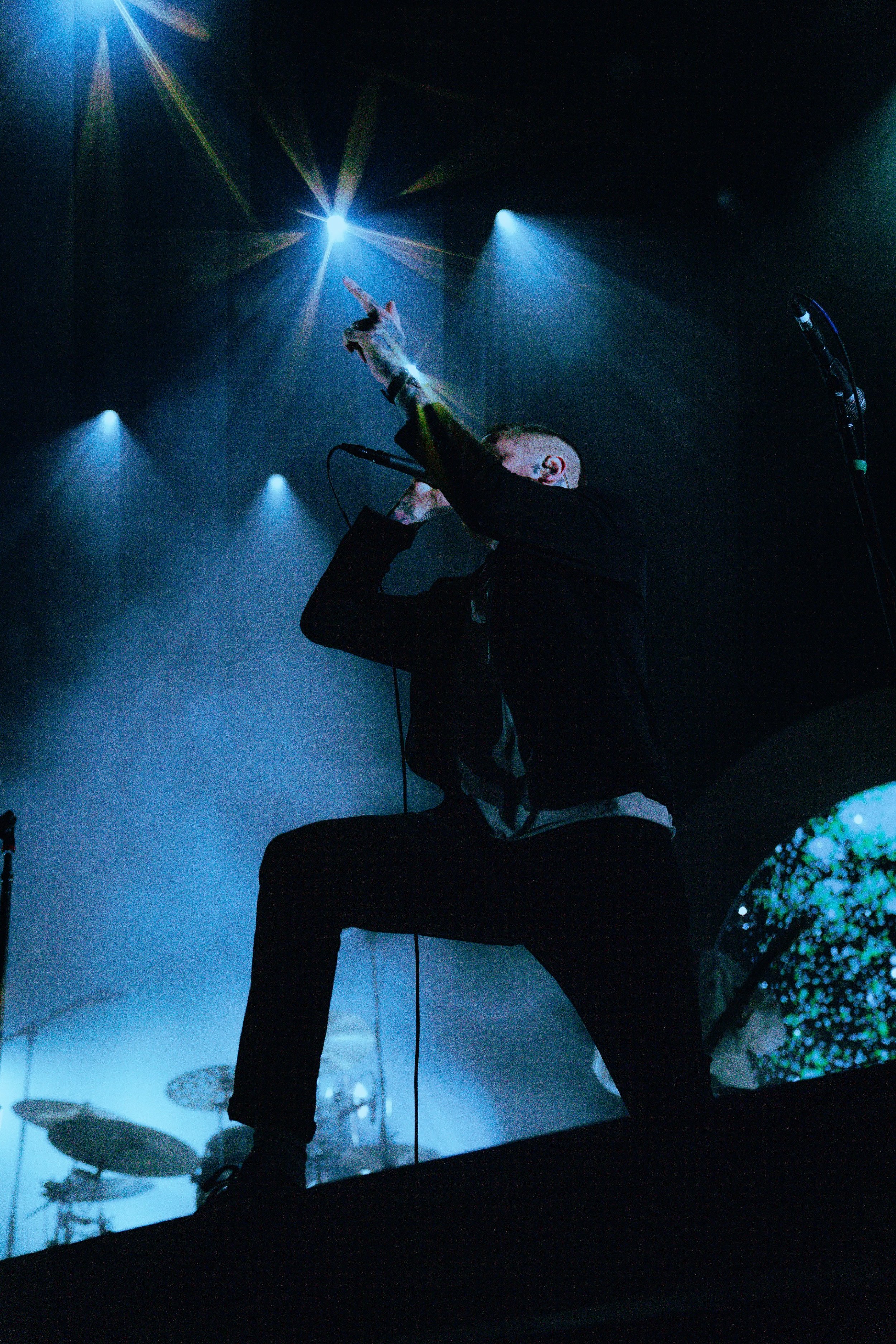 A singer on stage performs with a microphone, illuminated by blue and green stage lights, with a dimly lit drum set in the background.