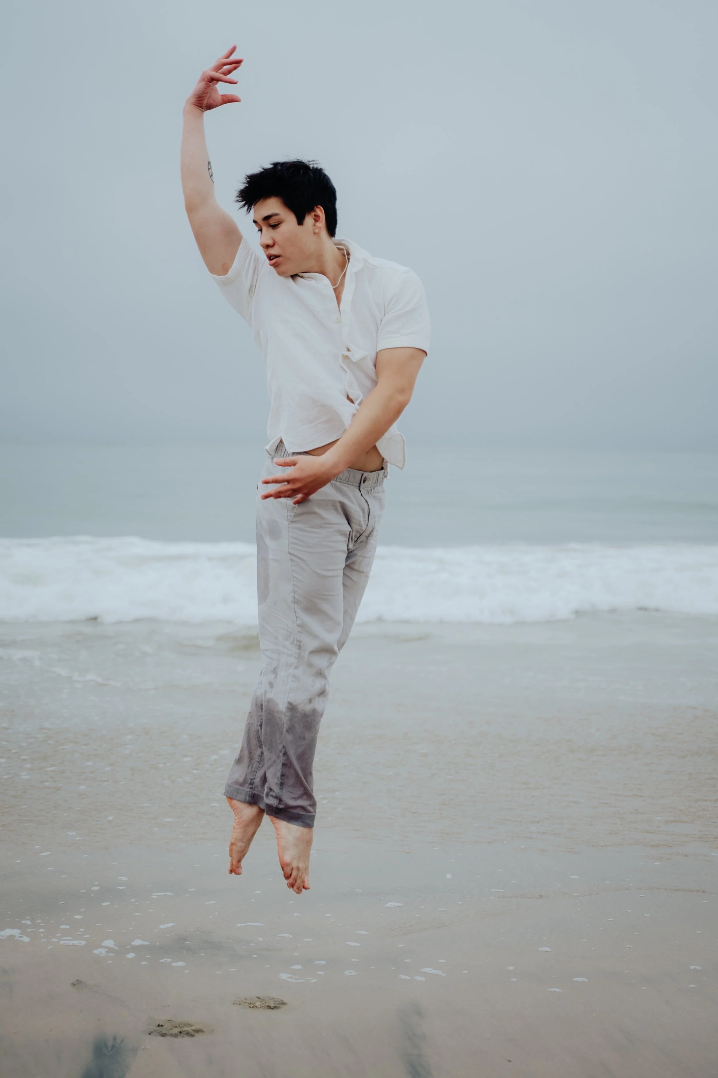 Person with short dark hair wearing a white shirt and light pants, jumping in the air at the beach with water and sand visible, overcast sky background.