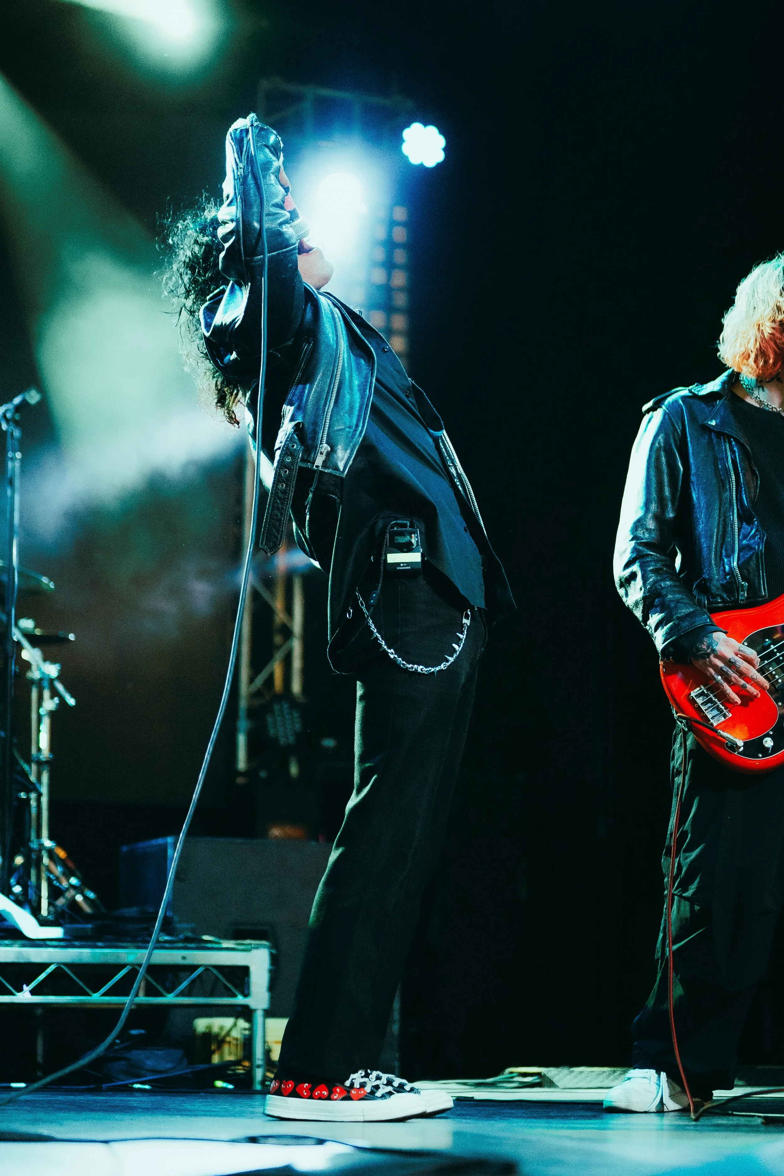 Close-up of a musician on stage, wearing a black leather jacket, black pants with chain details, and black and white sneakers with red heart patterns. The musician is holding a microphone and standing near music equipment, with stage lights in the ba