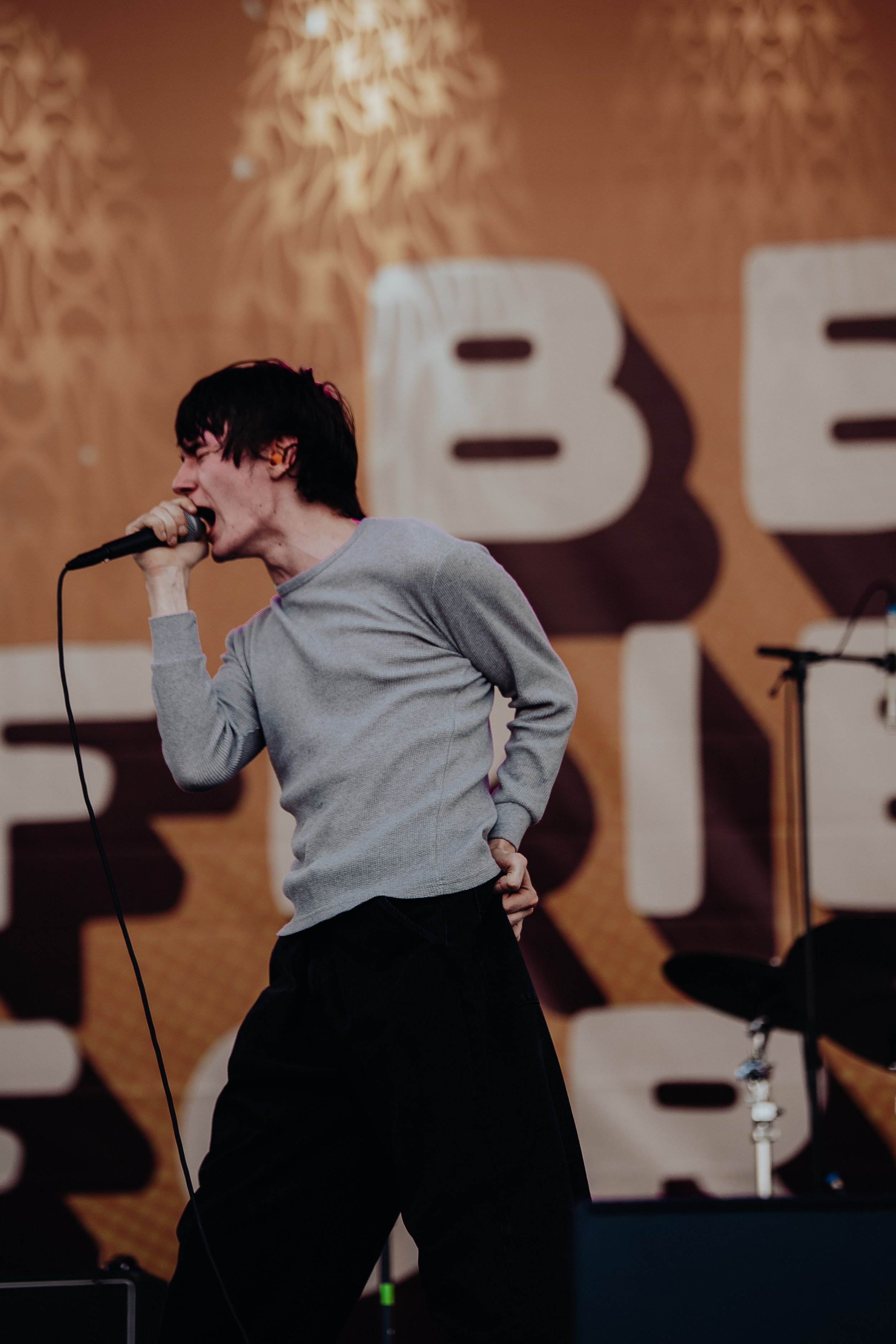 A male singer passionately performs on stage, holding a microphone with his right hand and his left hand behind his back. He has dark hair, is wearing a gray long-sleeve shirt and black pants. The background features a large, blurred decorative sign 