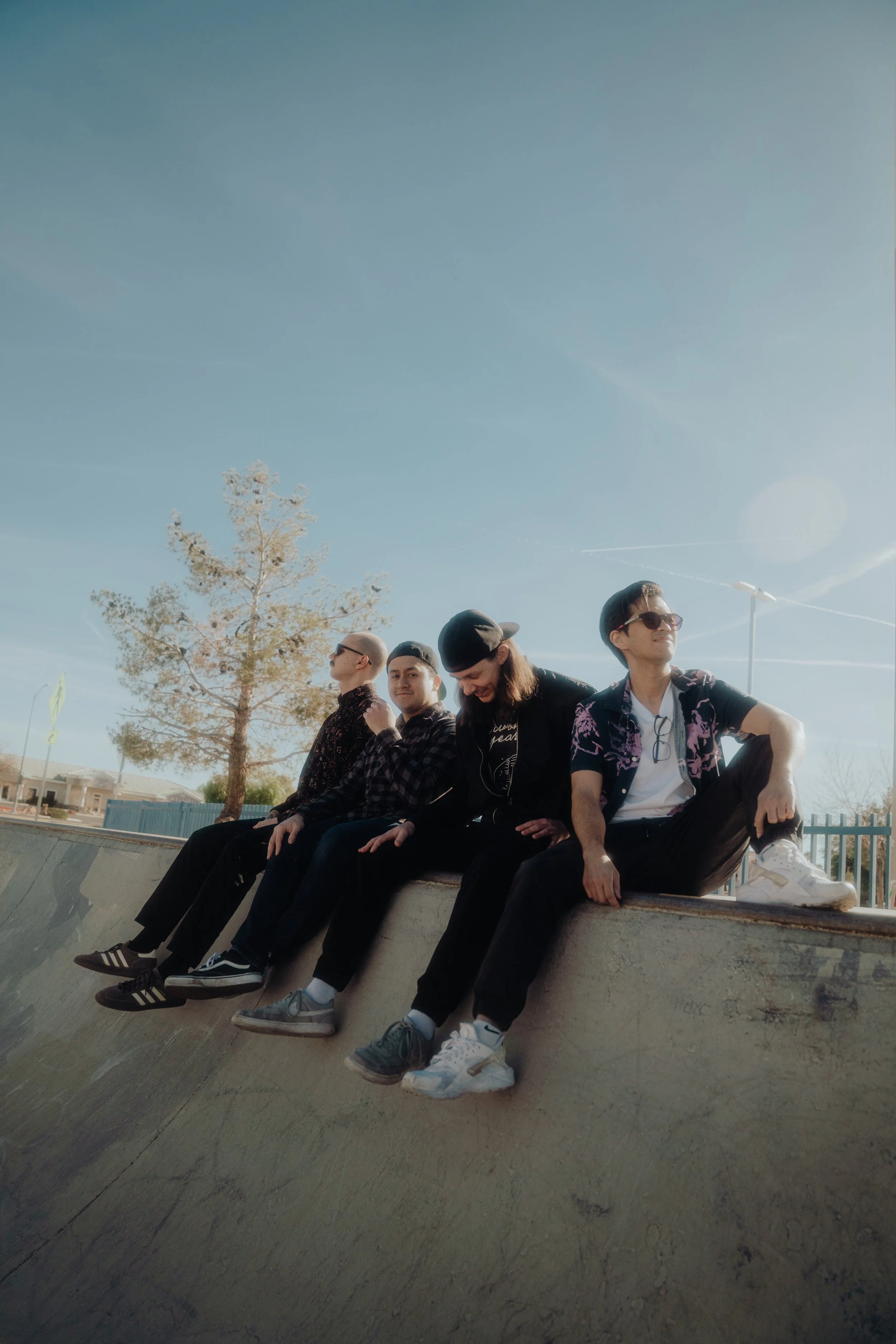 Five young men sitting together on a skateboard park ledge under a clear blue sky, some wearing sunglasses, with a tree and urban surroundings in the background.