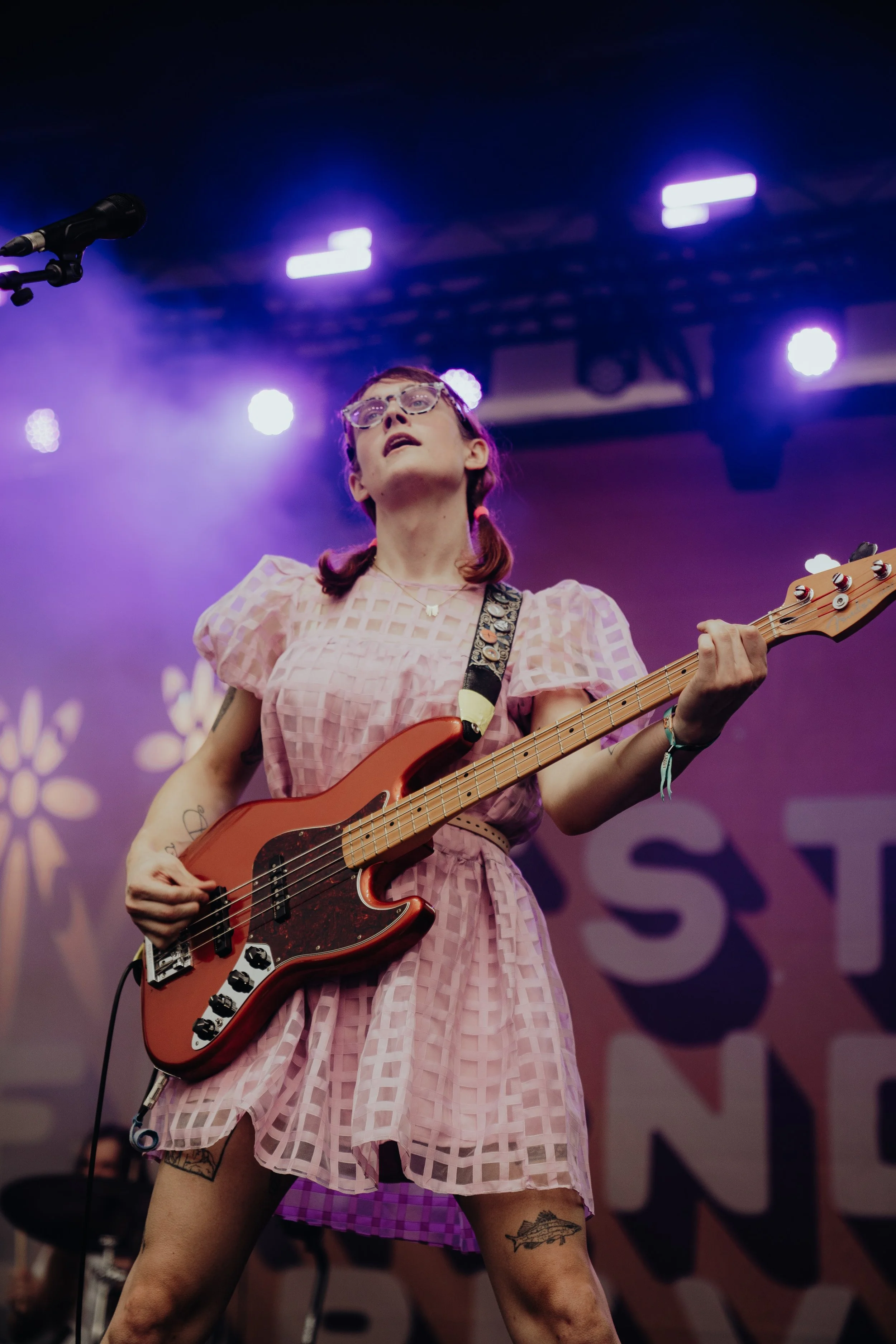 A woman in a pink checkered dress playing an orange electric bass guitar on stage with purple lighting.