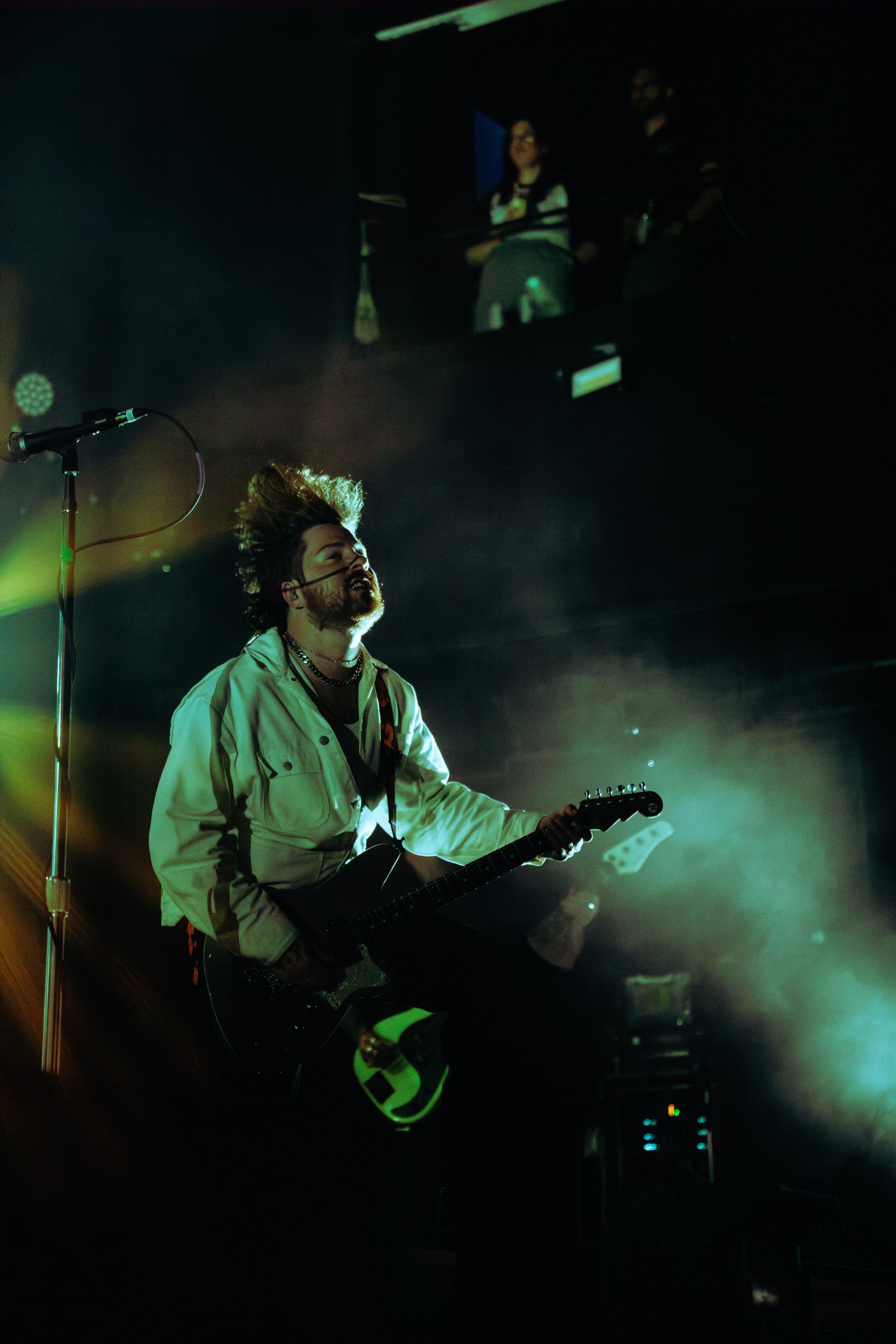 A male musician with a microphone headset and a beard playing an electric guitar on stage, with a dark background and greenish stage lighting. There is a large screen behind him showing two women and a man.