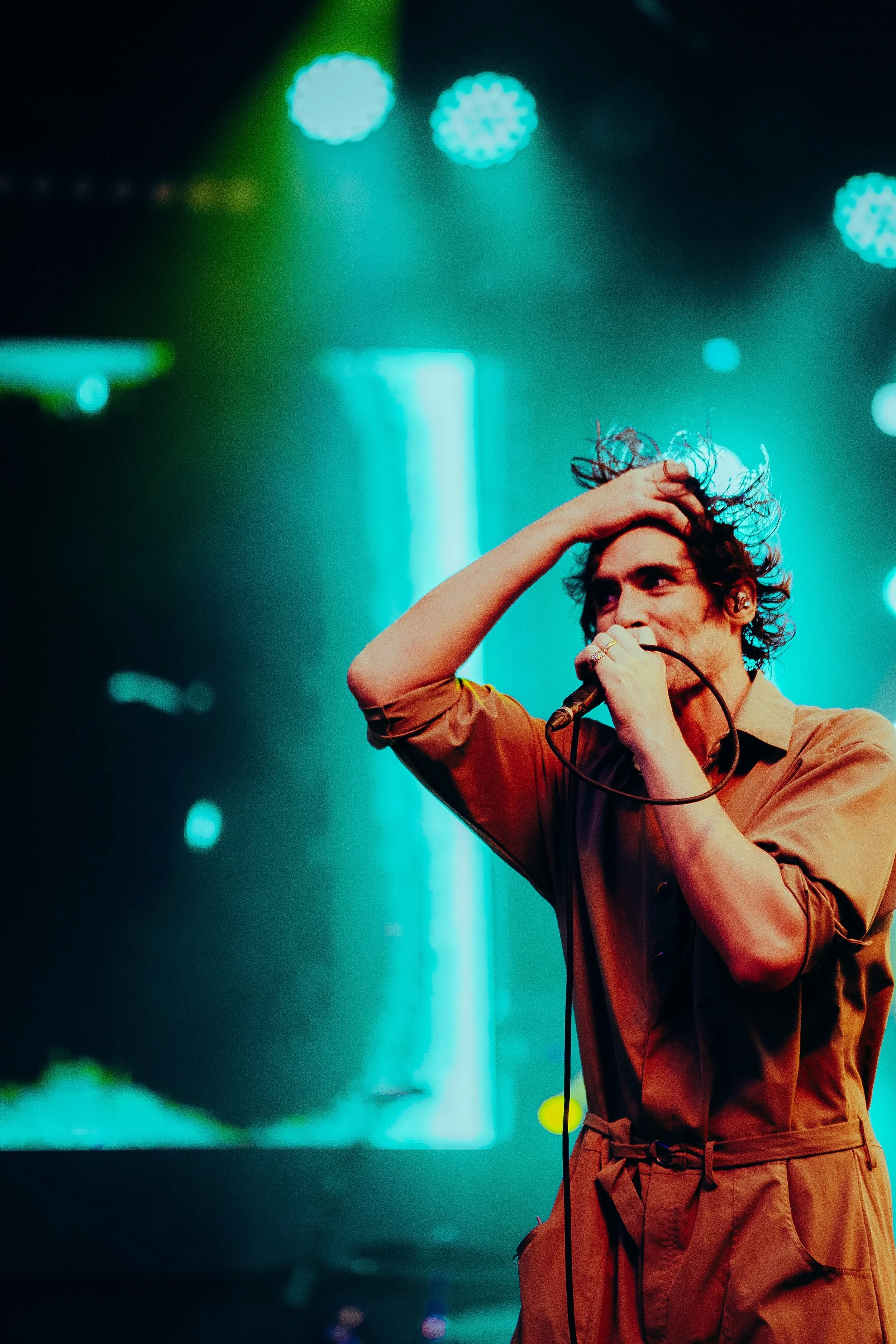 A person with curly hair on stage holding a microphone, with stage lights in the background creating an atmospheric concert scene.