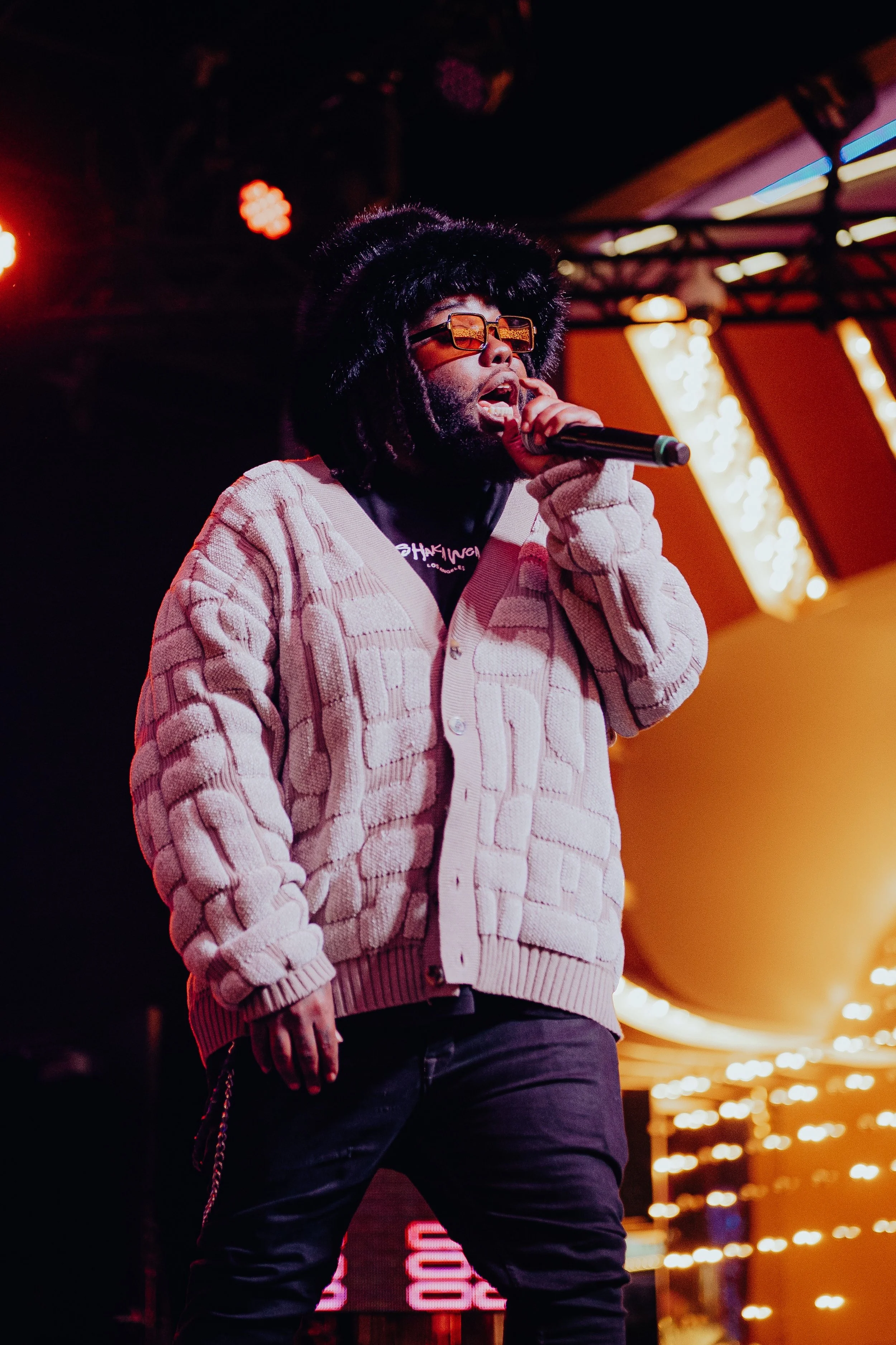 A male performer wearing a beige knit cardigan, black sunglasses, and a large fur hat, singing into a microphone on stage with colorful lights and a neon sign in the background.