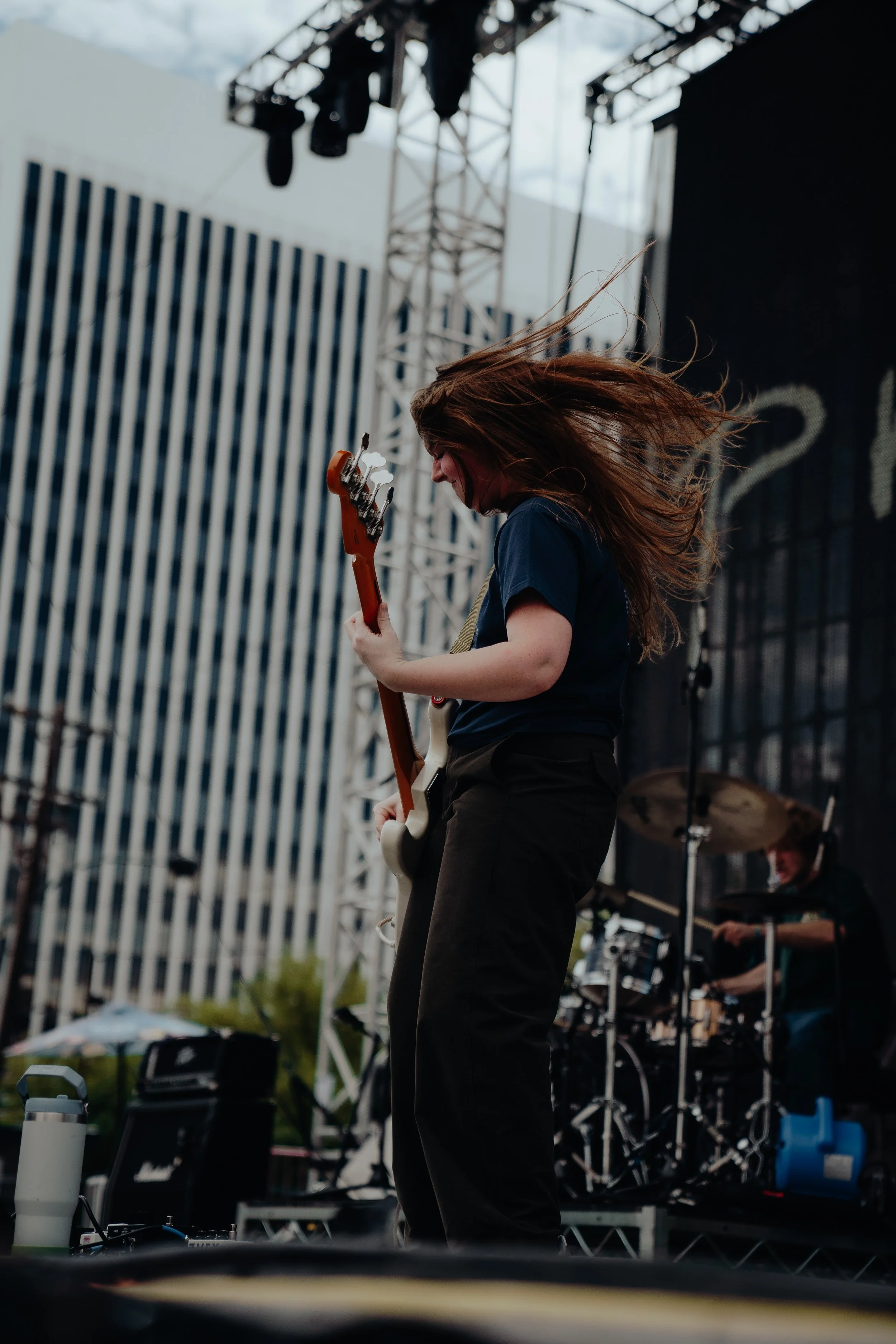 A woman with long hair playing an electric guitar on stage at an outdoor concert. She appears to be enjoying the music, and there is a drummer in the background. The setting is urban with modern buildings and a stage built with metal scaffolding.