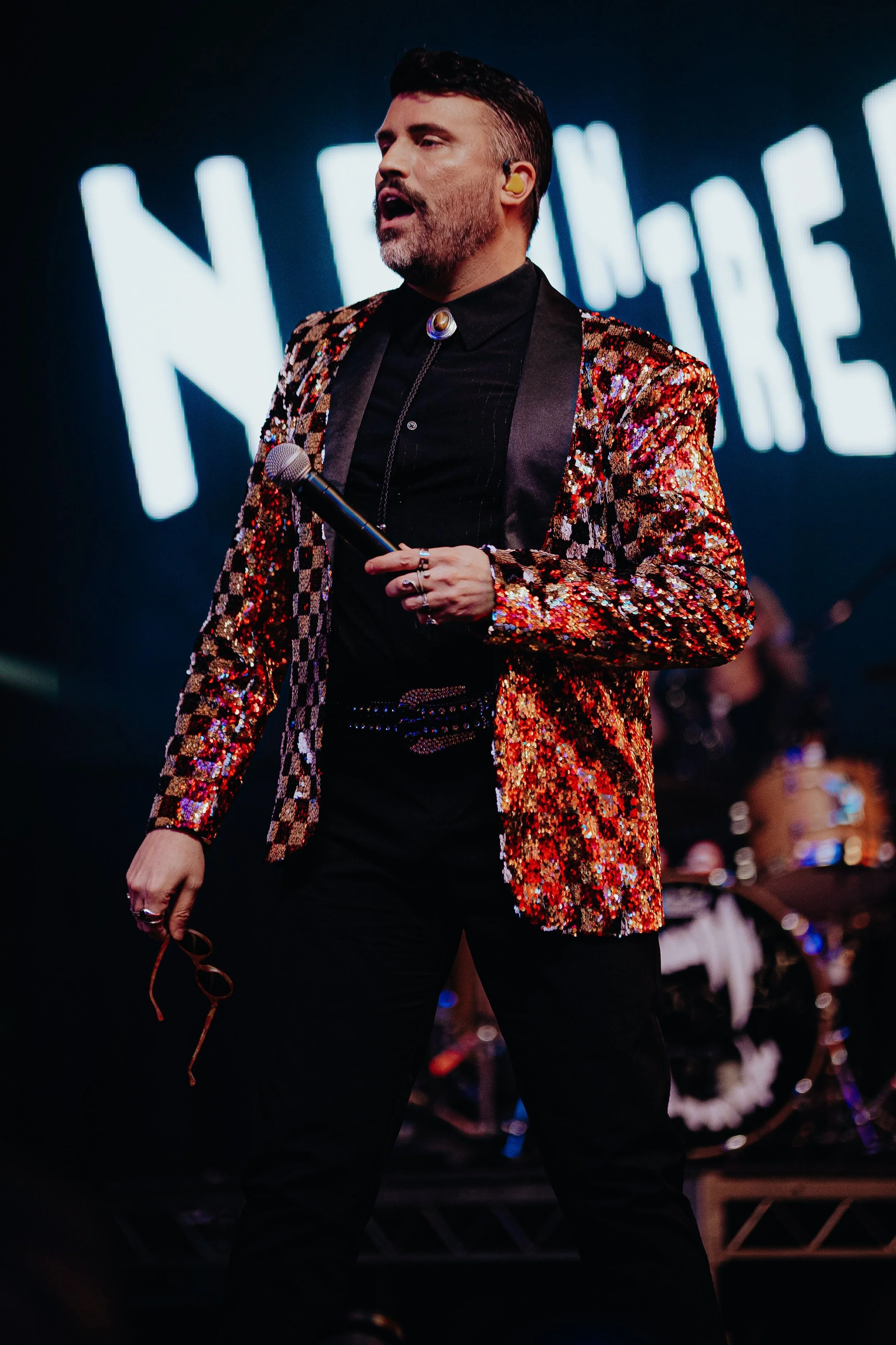 A man with a beard and short dark hair performing on stage, wearing a glittery, multicolored blazer, black shirt, and black pants, holding a microphone and glasses.
