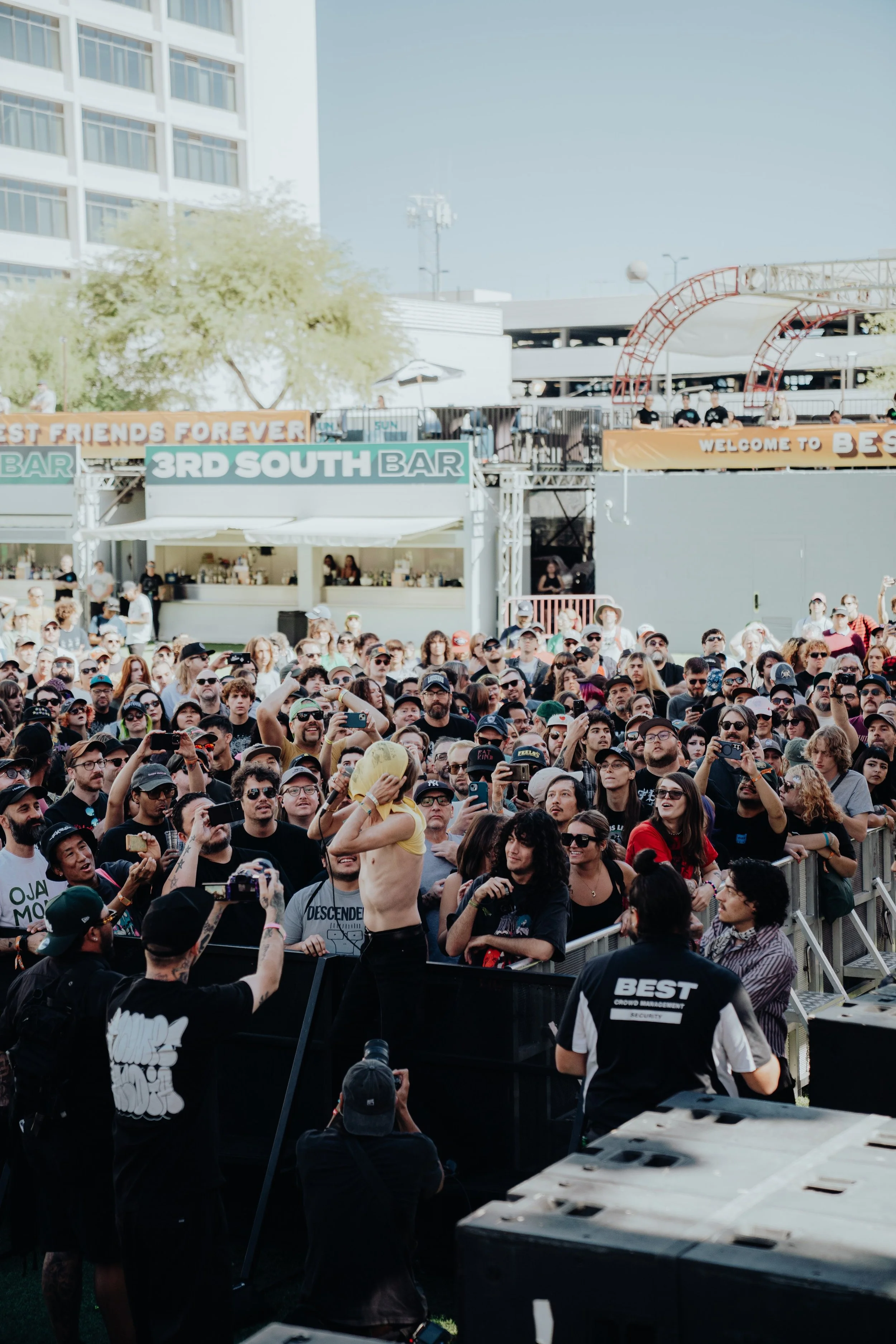 A large group of people attending an outdoor event, with some people taking photos and videos. There are signs for a bar and a stage in the background, along with a security guard in the foreground.