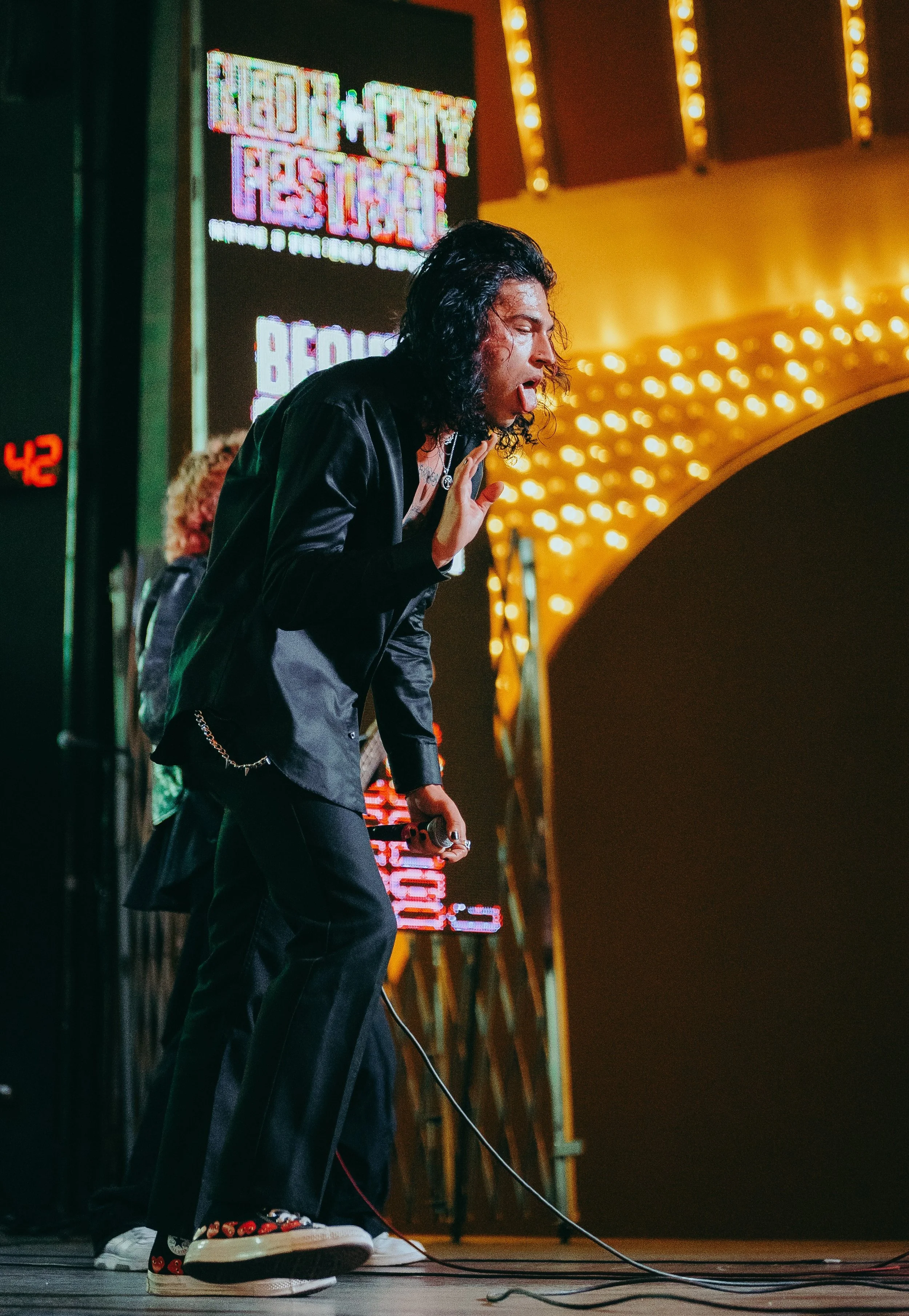 A young man with long black curly hair wearing a black jacket, black pants, and sneakers with a colorful heart pattern, holding a microphone, performing on stage at night. Bright signs and string lights are visible in the background.