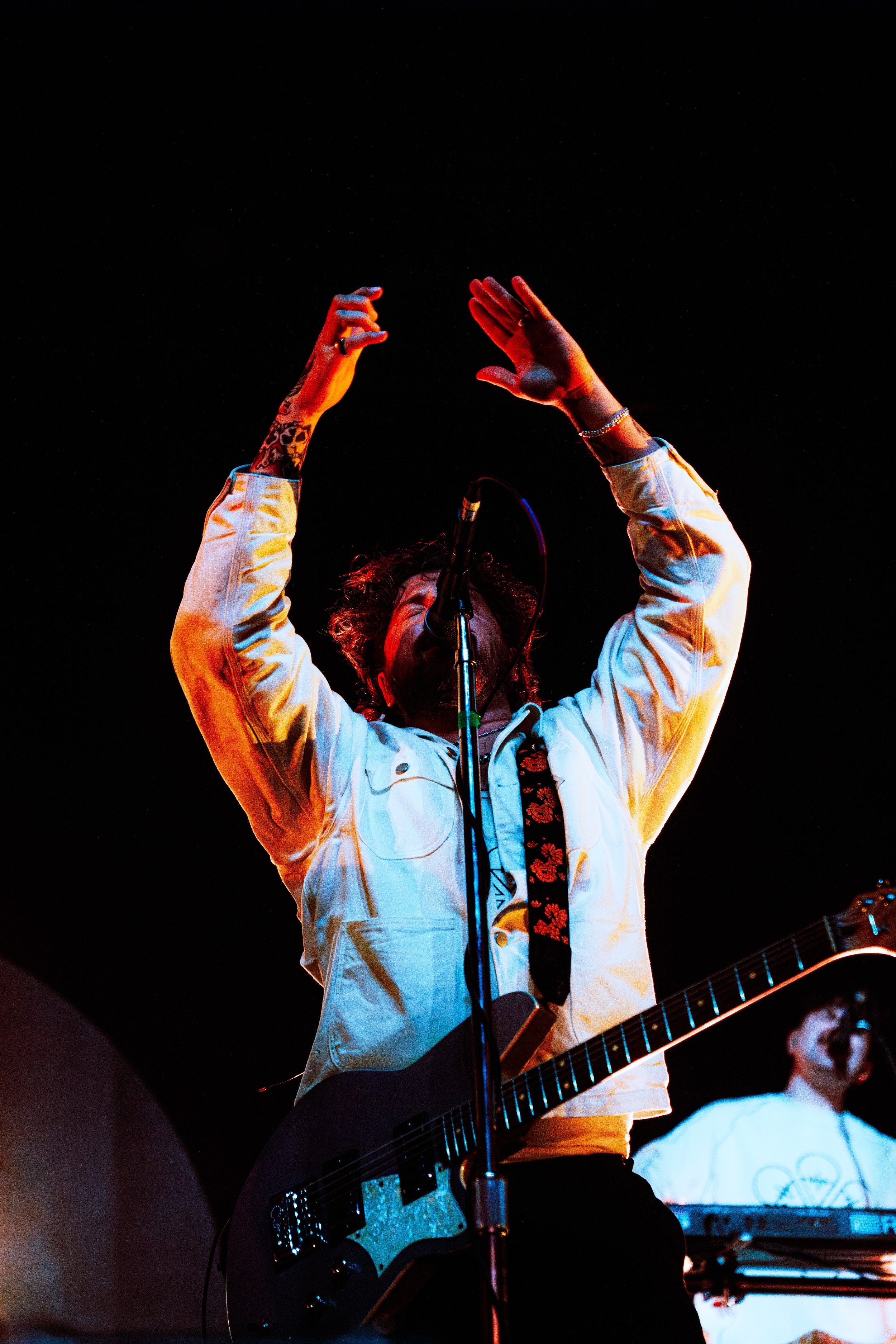 Person with curly hair raising hands, wearing a white jacket with floral embroidery, playing guitar during a performance, with another musician in the background wearing glasses and a white shirt.