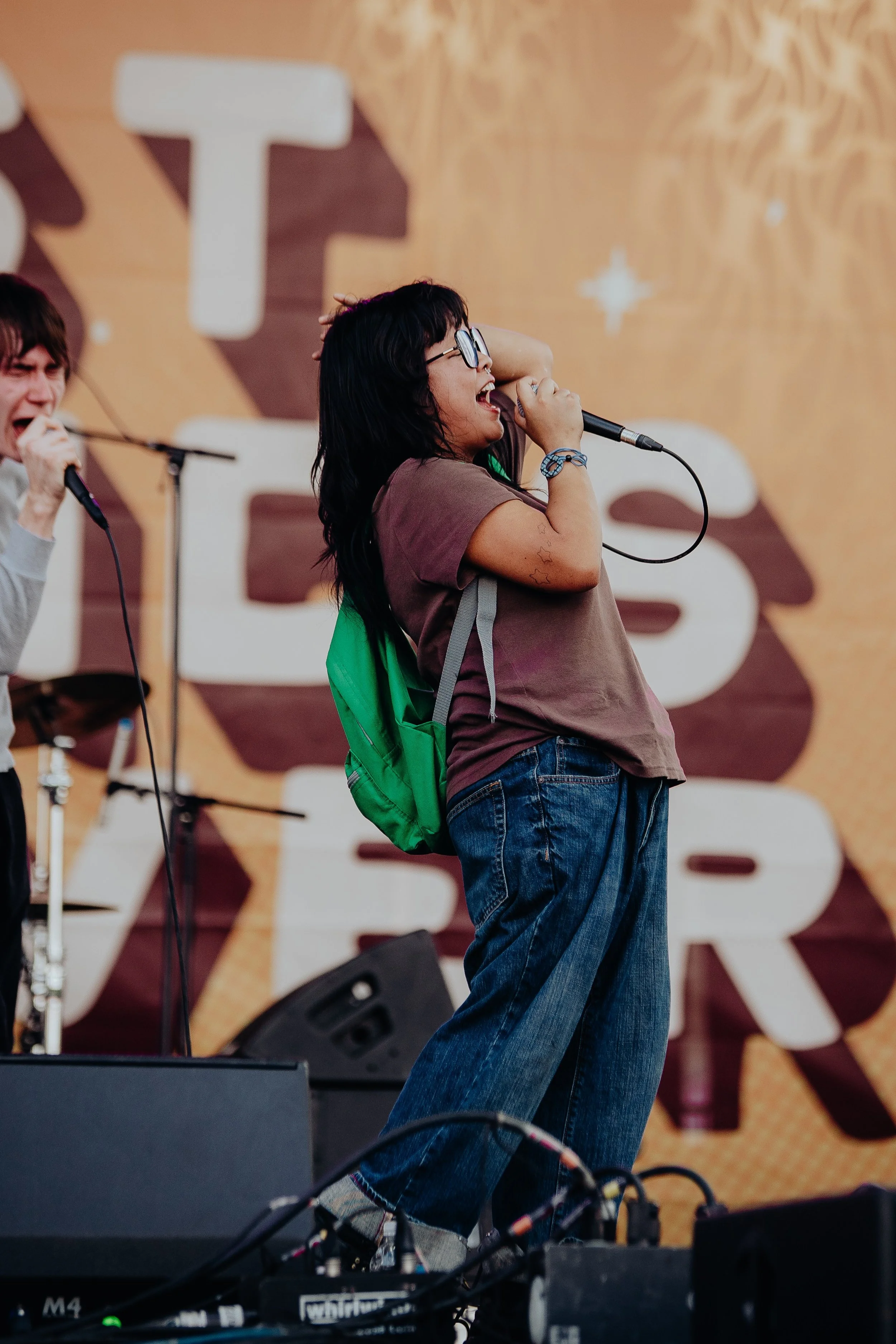 A woman with black hair, glasses, and tattoos on her arm is singing into a microphone on stage. She is wearing a brown shirt and jeans, and has a green backpack. Another person is partially visible on the left side, also holding a microphone. The bac