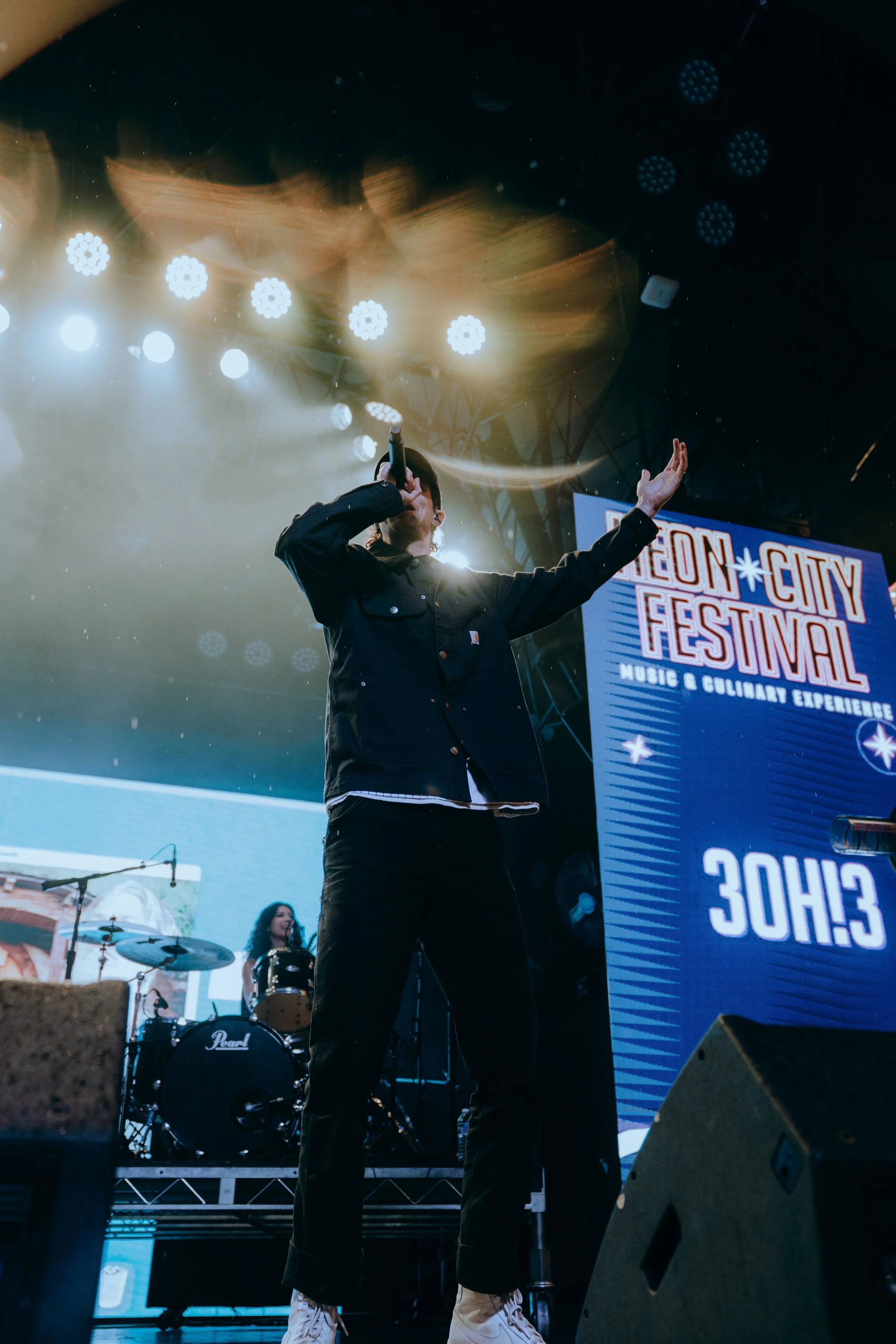 A singer performing on stage at the Neon City Festival with a band in the background, stage lighting, and a large festival sign.