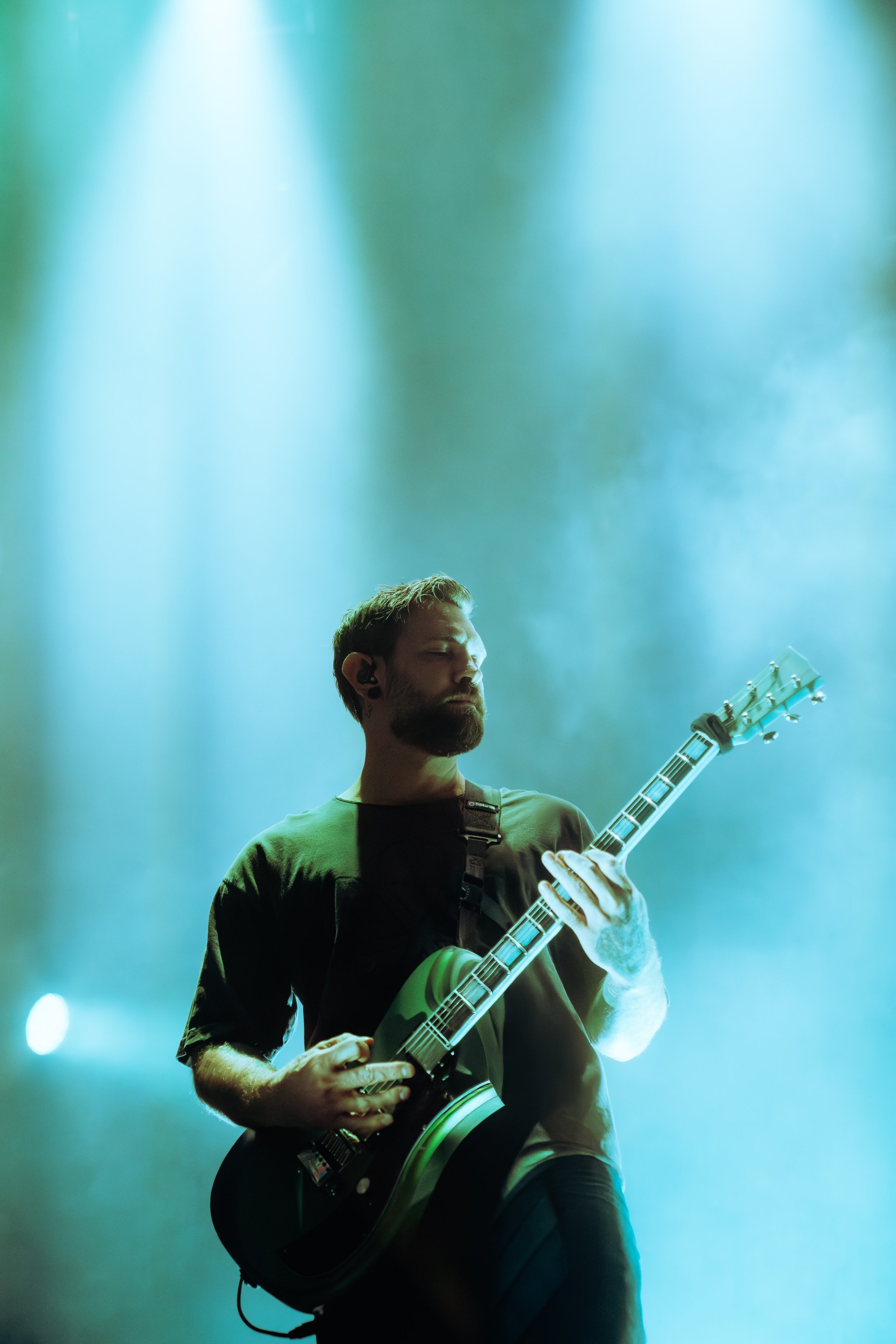 A male musician playing an electric guitar on stage, with blue and green stage lights illuminating the background.