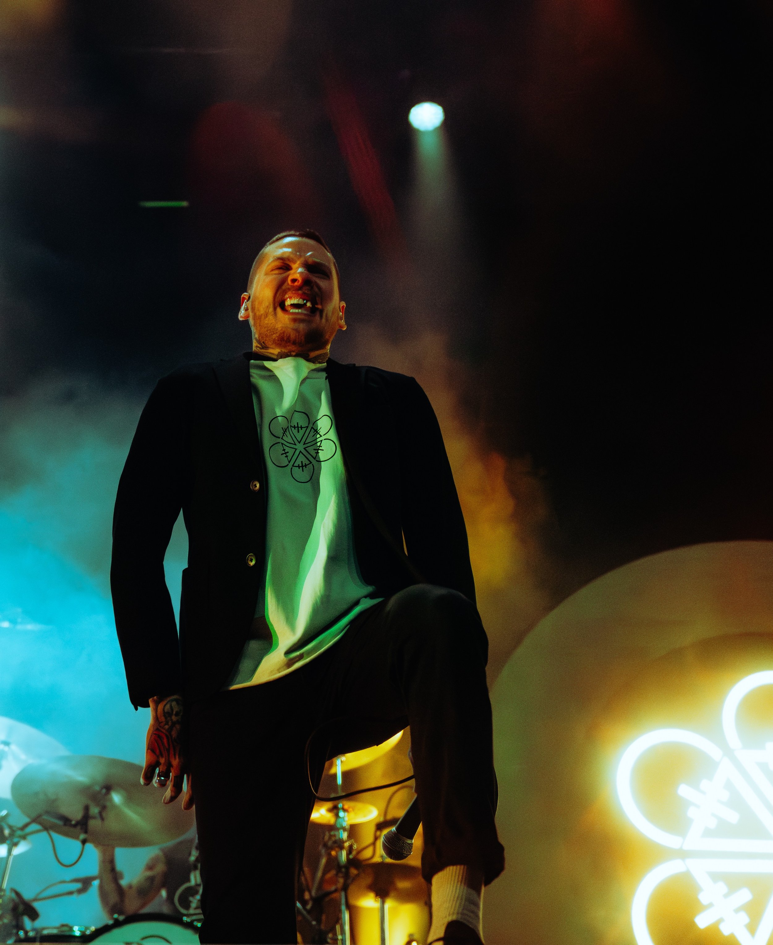 A man with a beard and tattoos on his hands, wearing a black blazer and white shirt, laughing on stage during a performance. The background features colorful lighting and musical equipment.