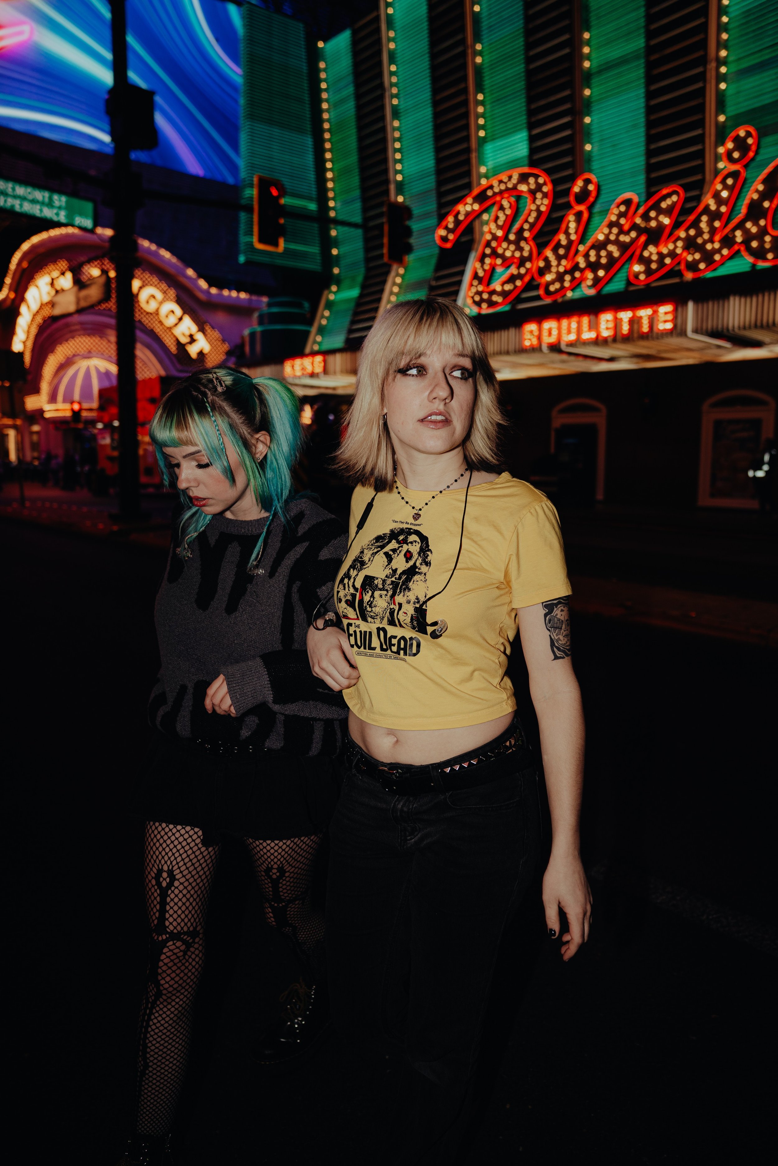 Two young women walking at night in front of a brightly lit neon sign that says 'Binion's'. One woman has blonde hair, wearing a yellow 'The Evil Dead' T-shirt and black pants. The other has green and blue hair, wearing a black and gray striped sweat