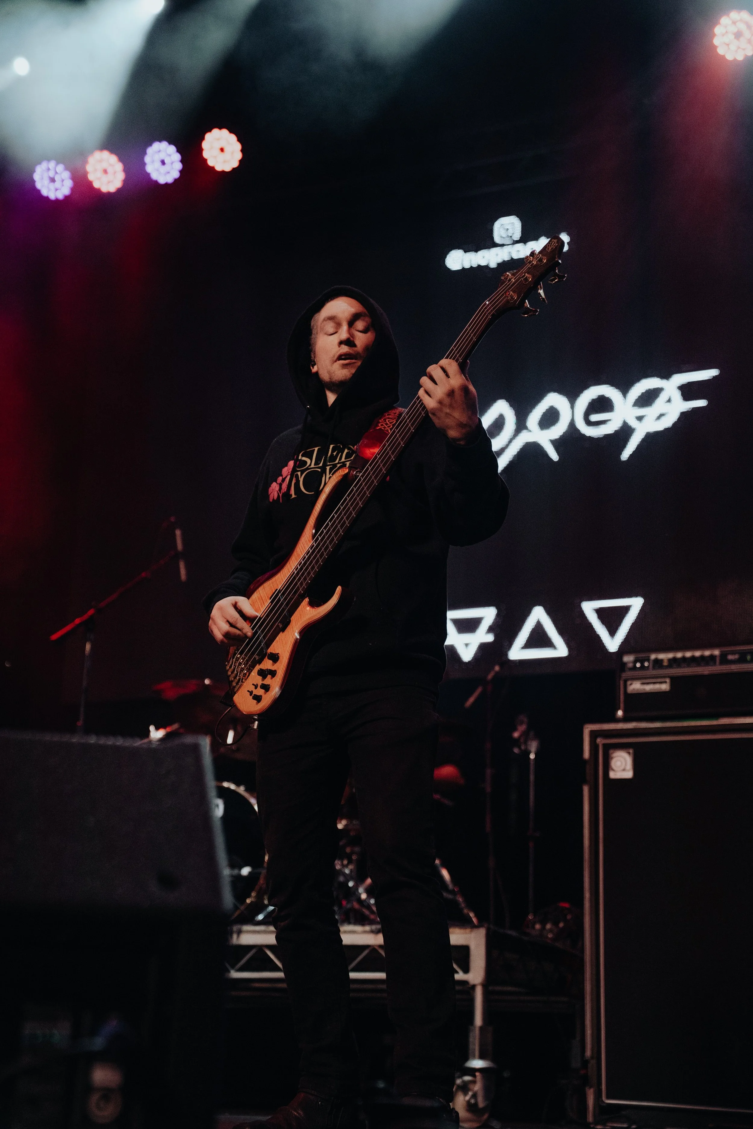 A man in a black hoodie playing a bass guitar on a stage with colorful lights and neon signs in the background.