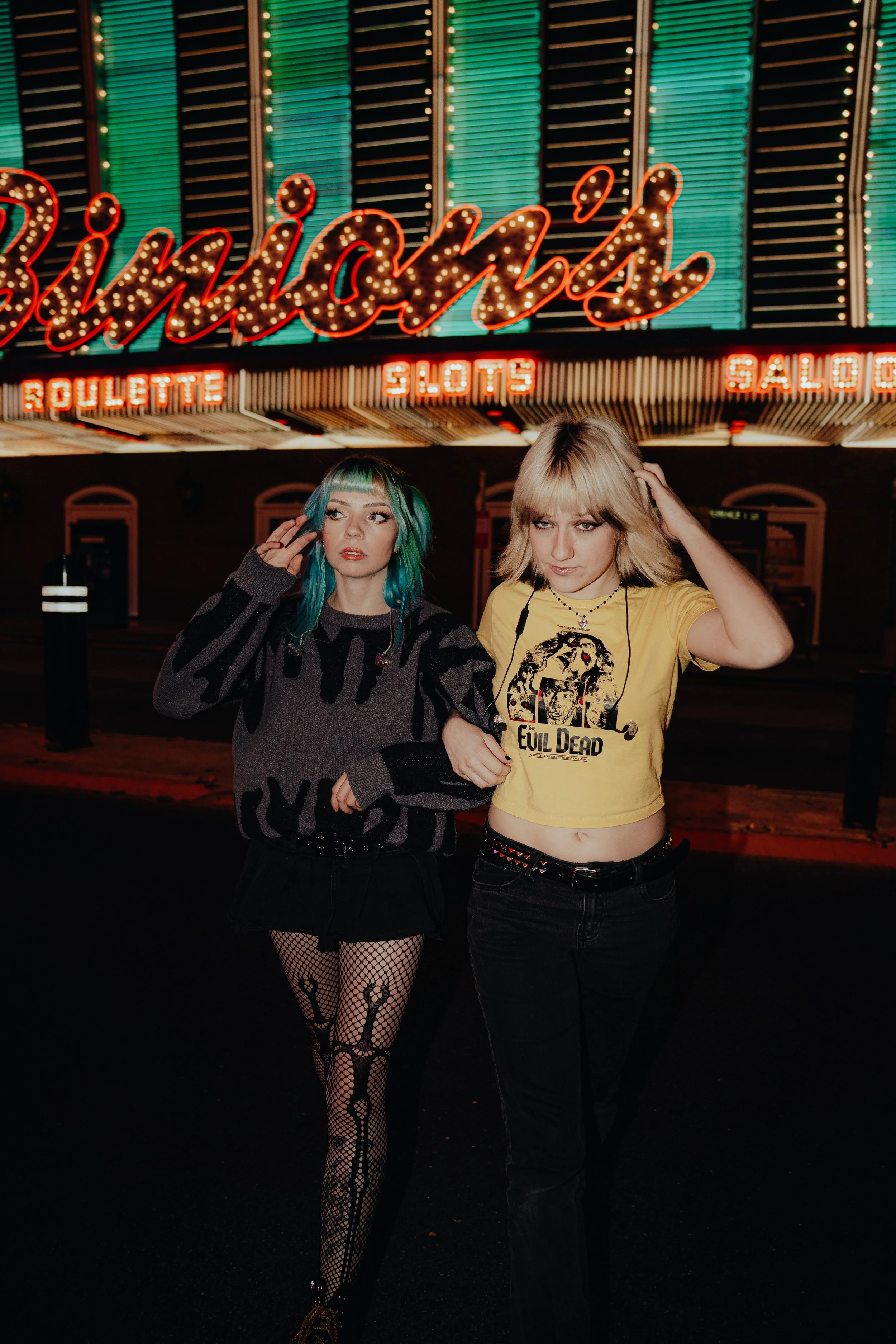 Two young women stand in front of a colorful neon sign that reads 'Bingo'. The woman on the left has teal and green hair, is wearing a black and gray oversized sweater, fishnet stockings with black skeleton designs, and is holding her hair. The woman