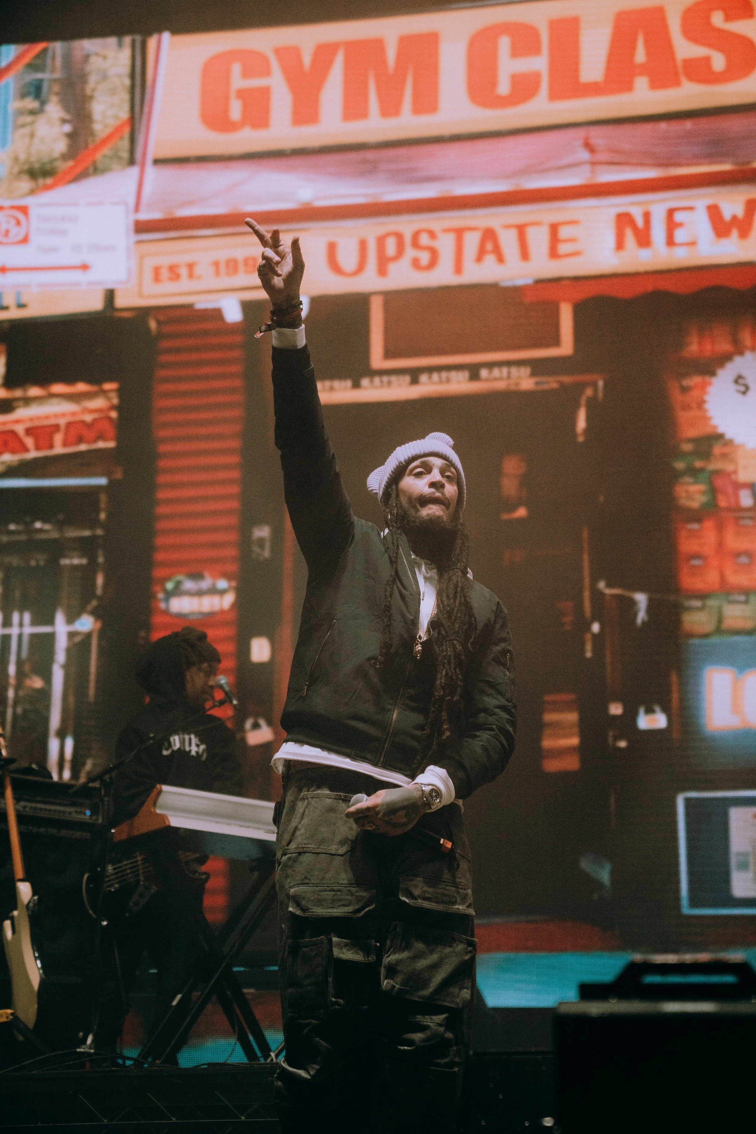 A man with long dreadlocks wearing a gray beanie, black jacket, and cargo pants, raising his right hand with two fingers in a peace sign, performing on stage with a keyboard player in the background and a city street scene displayed on a large screen