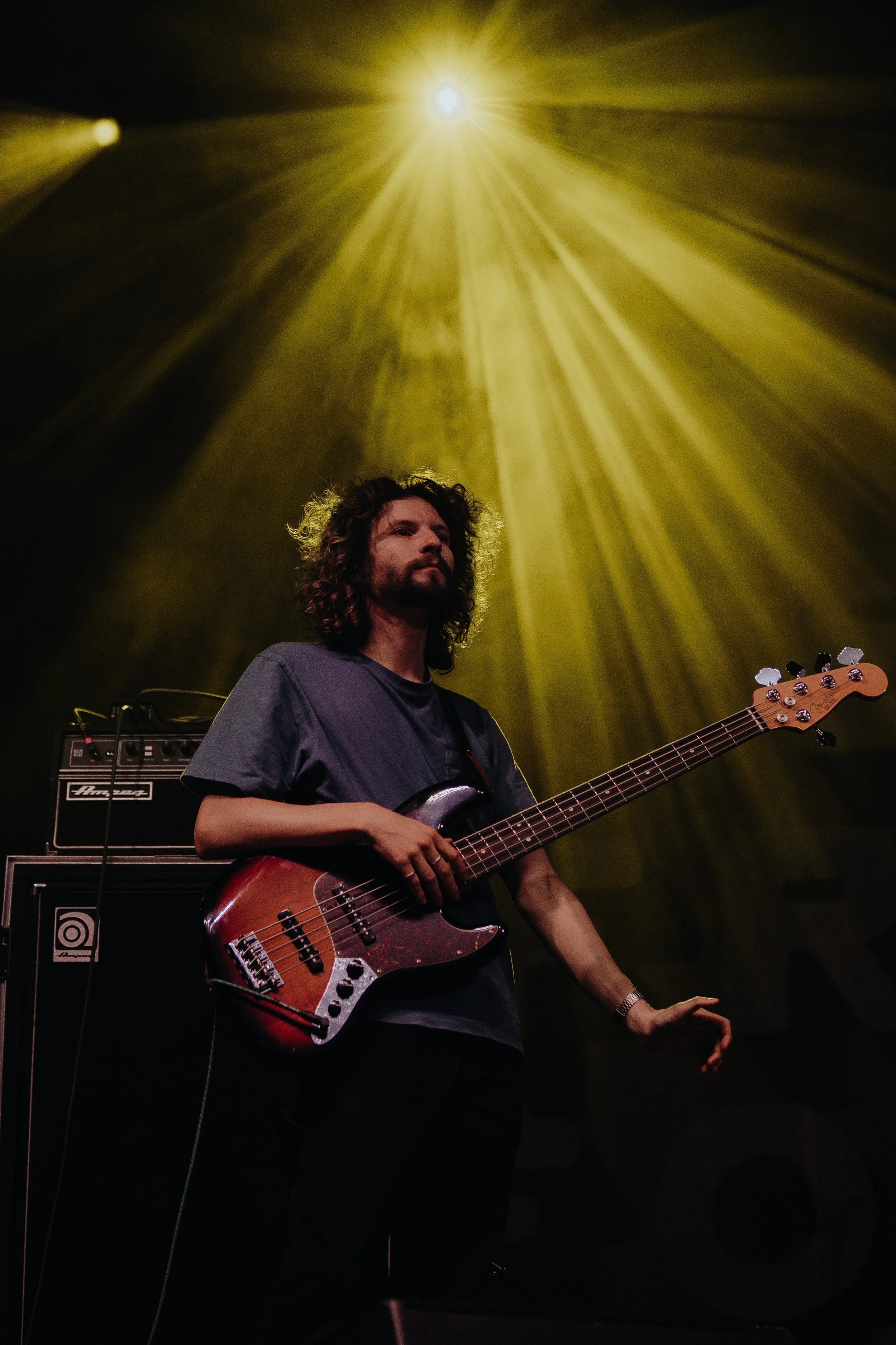 A man with curly hair and a beard playing an electric bass guitar on stage, with a large amplifier behind him and bright yellow stage lighting.