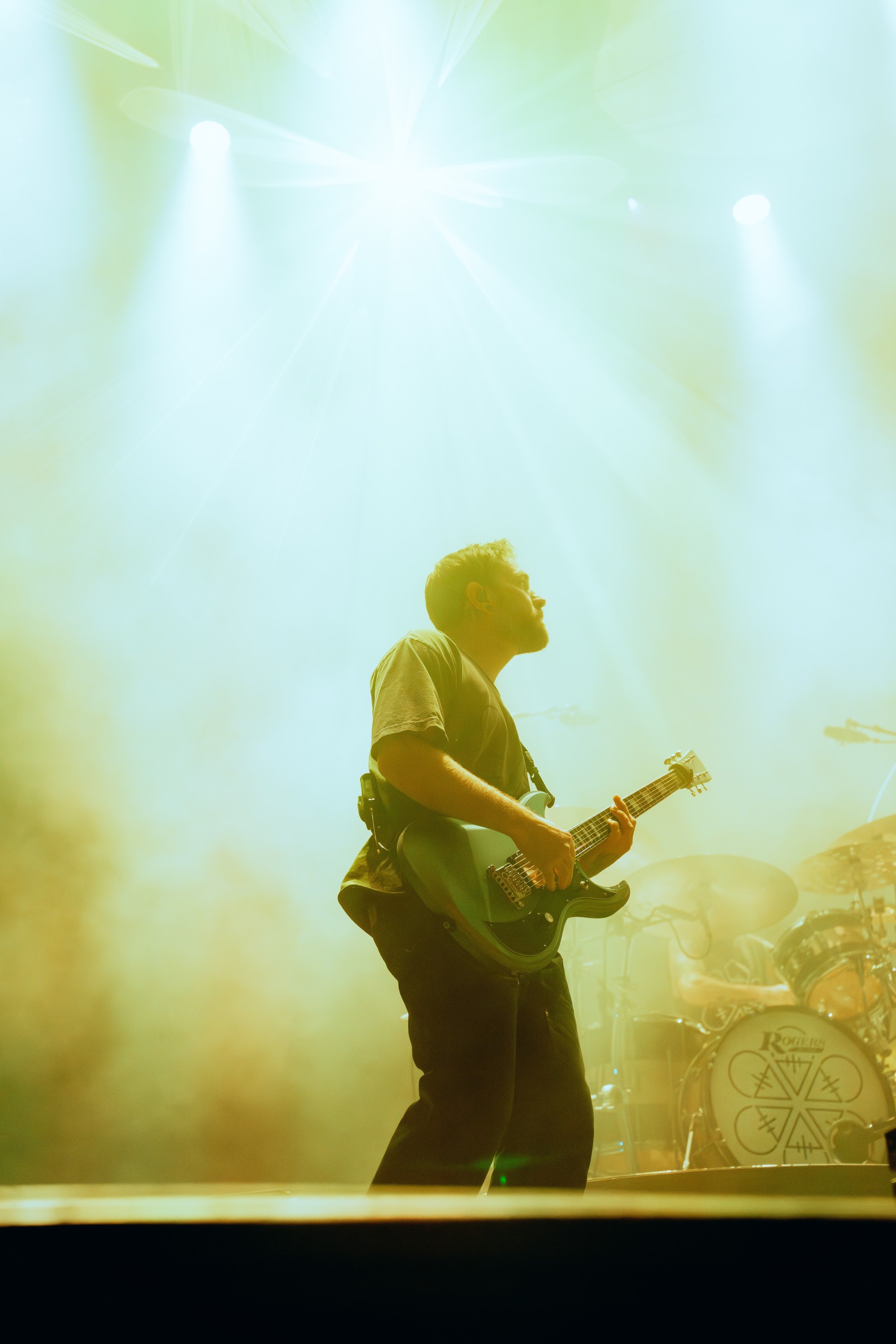 A male musician playing an electric guitar on stage, with a drum set in the background and stage lights shining down.