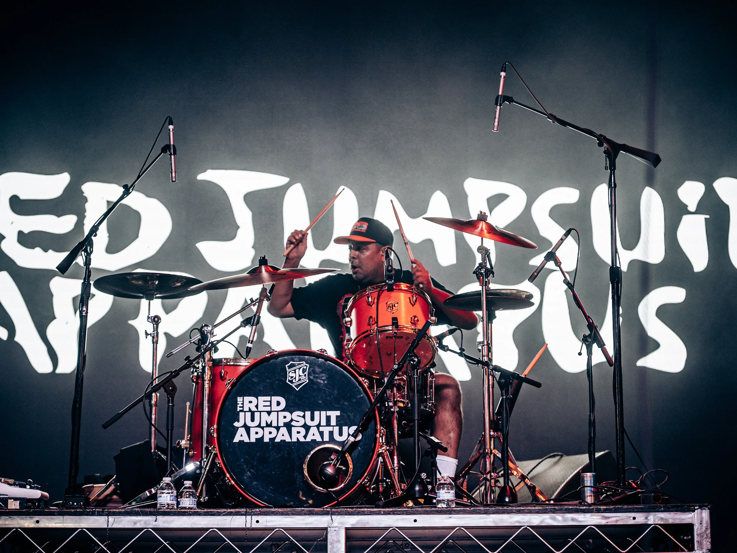 Musician playing drums on stage with a backdrop displaying the text 'The Red Jumpsuit Apparatus' and microphones hanging above the drum set.